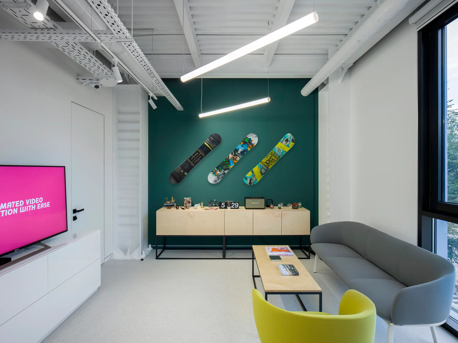 A modern office lounge featuring a grey sofa, a lime green armchair, and three snowboards mounted on a deep green accent wall.