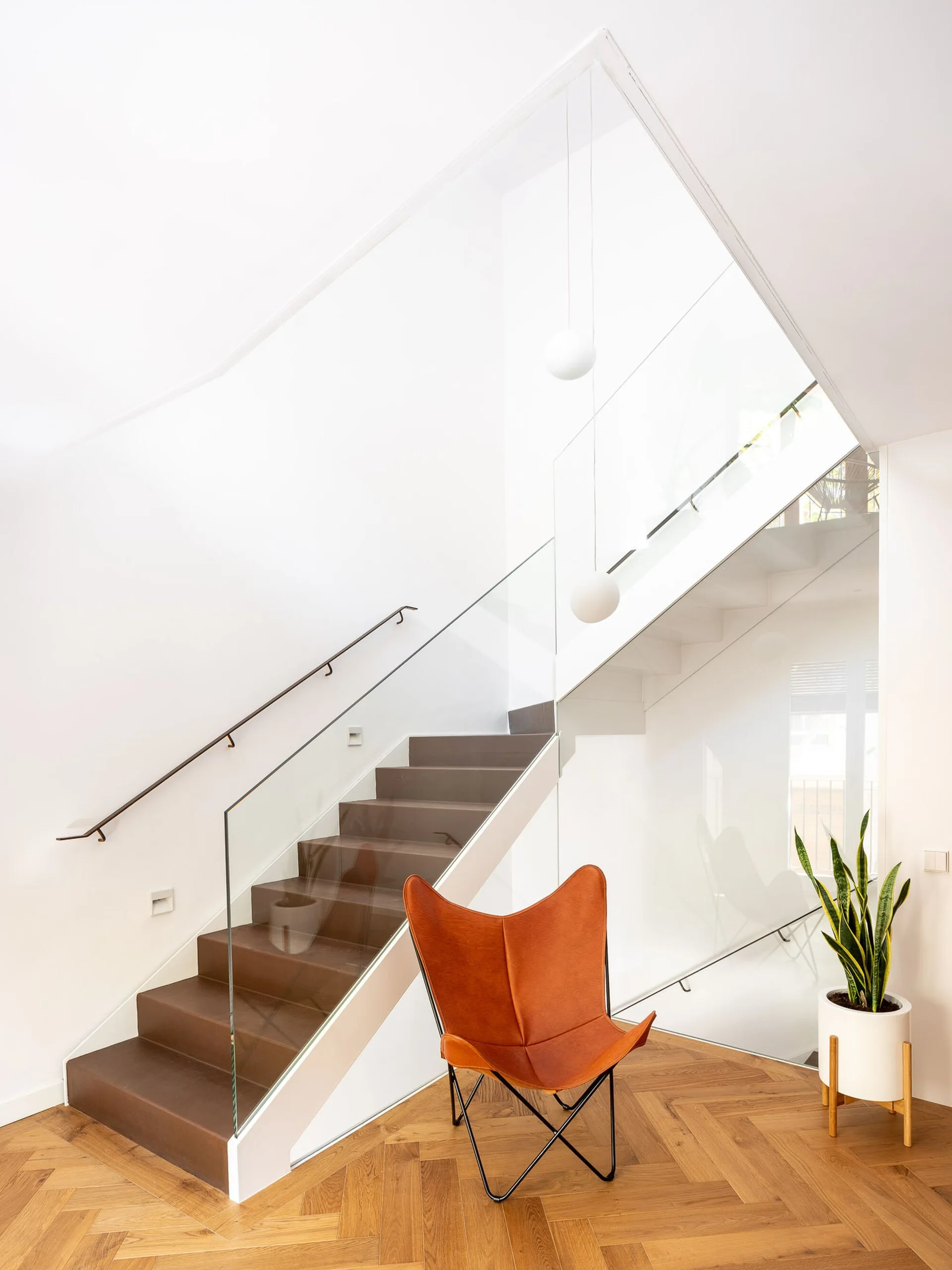 A modern staircase with a clear glass railing sits above a herringbone wood floor with a cognac leather butterfly chair.