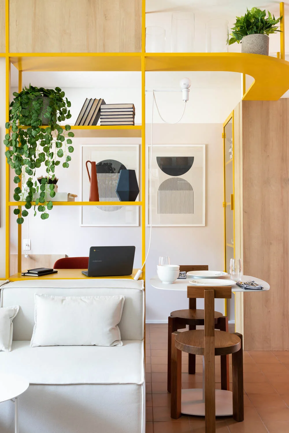 A bright yellow metal shelving unit divides a living area with a white sofa from a small dining table and wooden chairs.