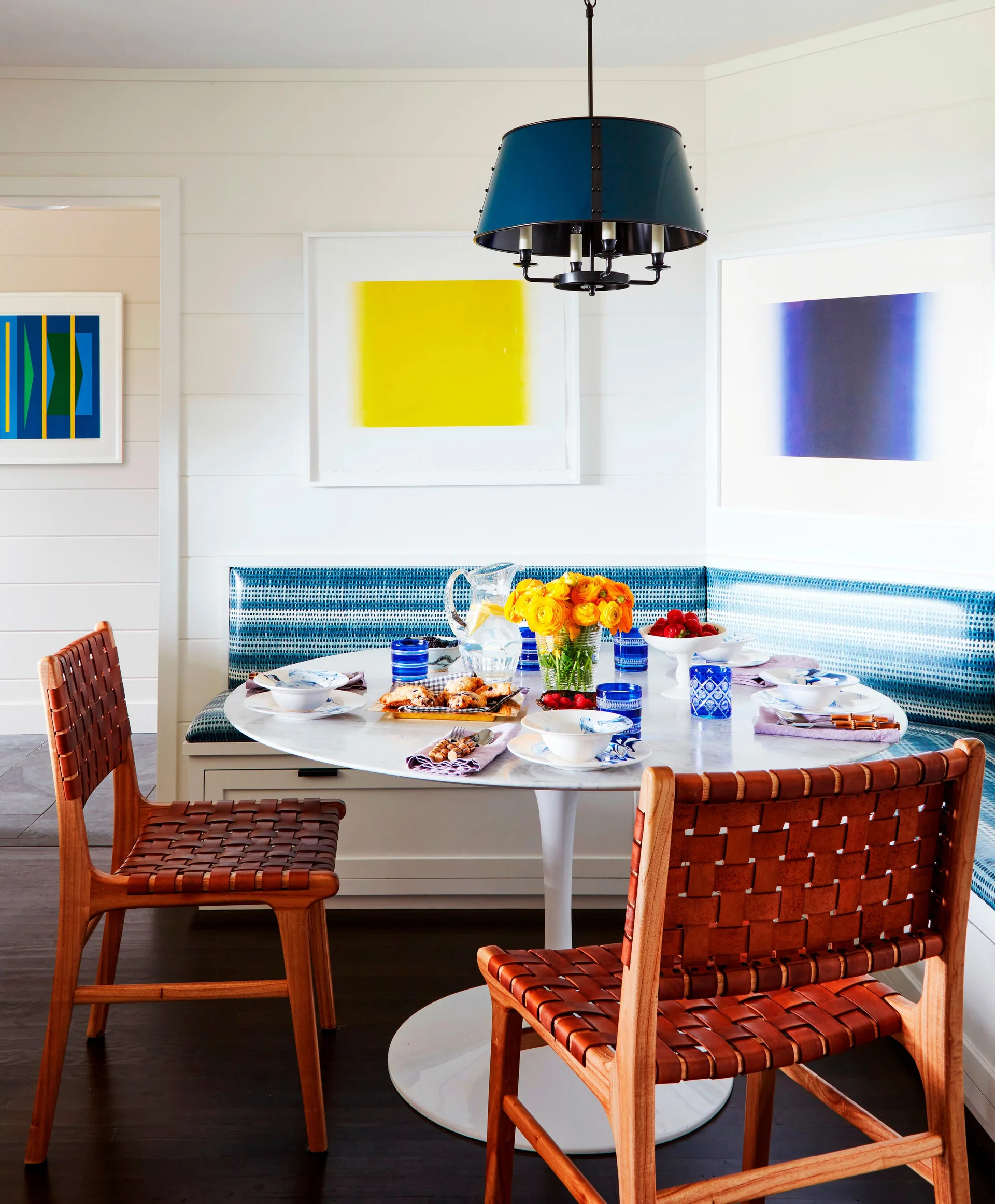 A bright dining nook features a white marble pedestal table, a built-in banquette with blue patterned upholstery, and two woven leather chairs.