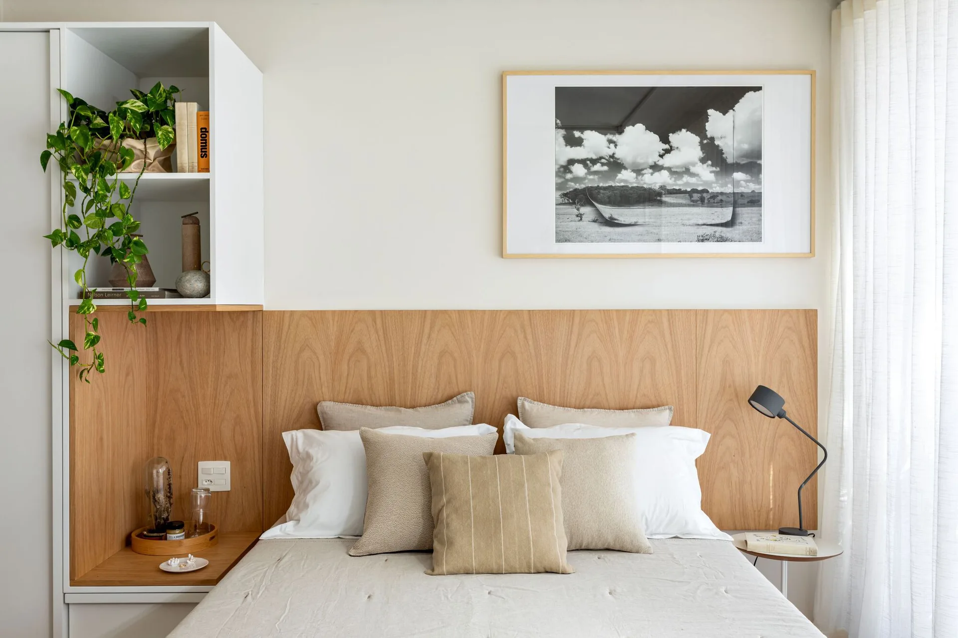 A minimalist bedroom features a light wood paneled headboard integrated with white shelving, neutral linen bedding, and a framed monochrome print.