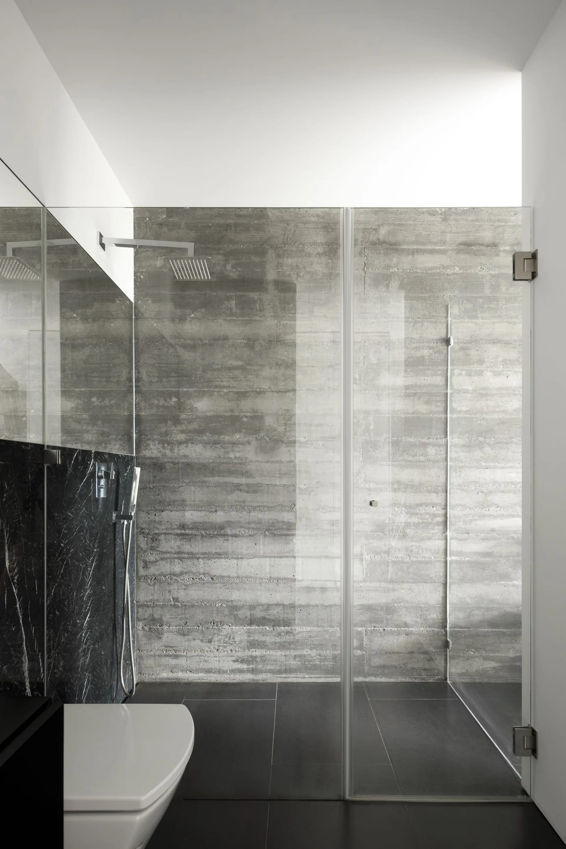 A minimalist bathroom features a glass-enclosed walk-in shower with board-formed concrete walls, a dark marble accent wall, and a white wall-mounted toilet.