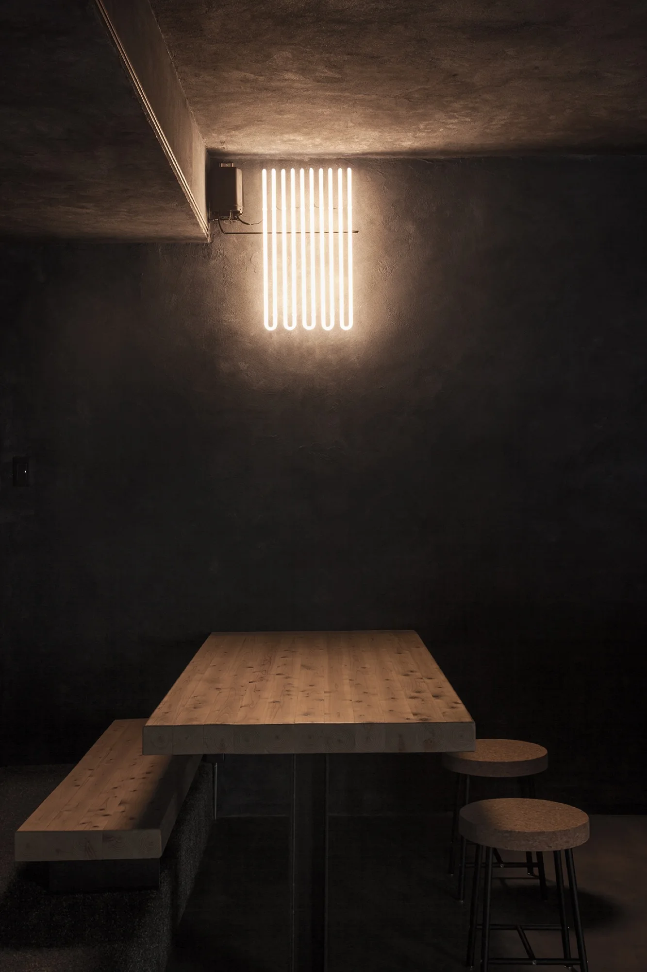 A minimalist dining corner features a light wood table and bench set against a dark textured wall beneath a glowing vertical tube light fixture.