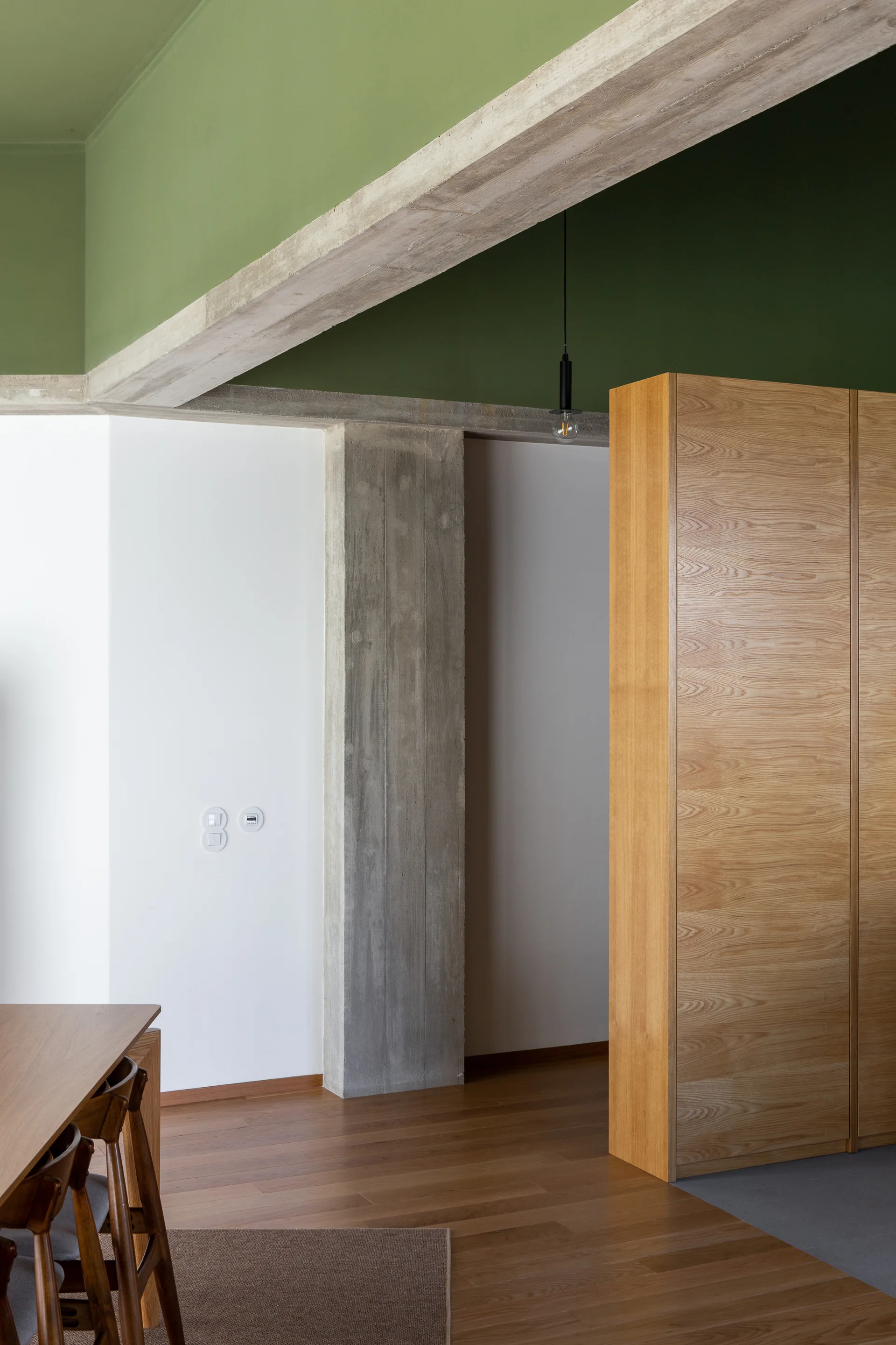 An interior space features green ceiling panels, exposed concrete beams and columns, a large wood cabinet, and a wooden dining set.