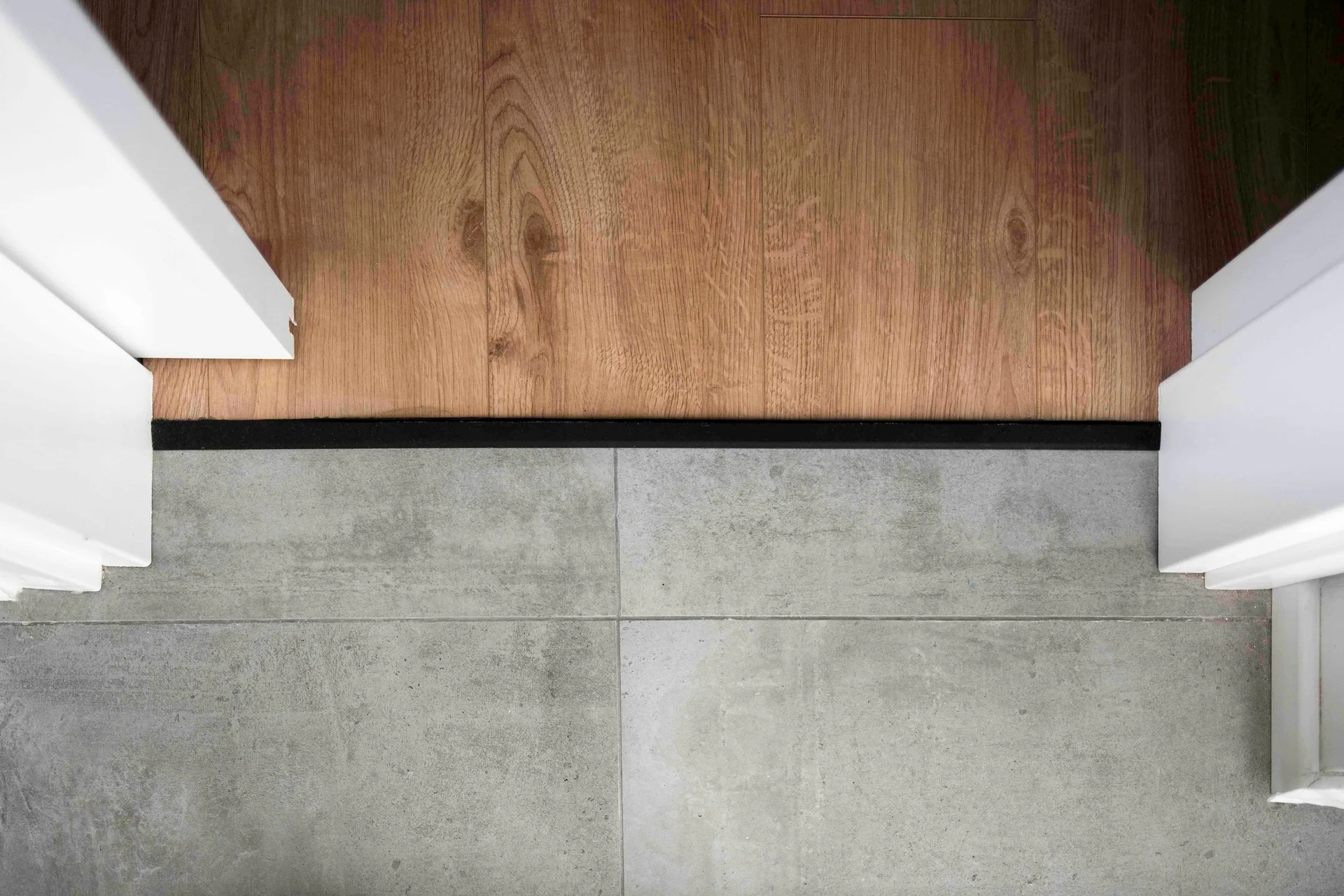 A top-down view shows a brown wood plank floor transitioning to a gray tile floor, separated by a thin black strip between white door frames.