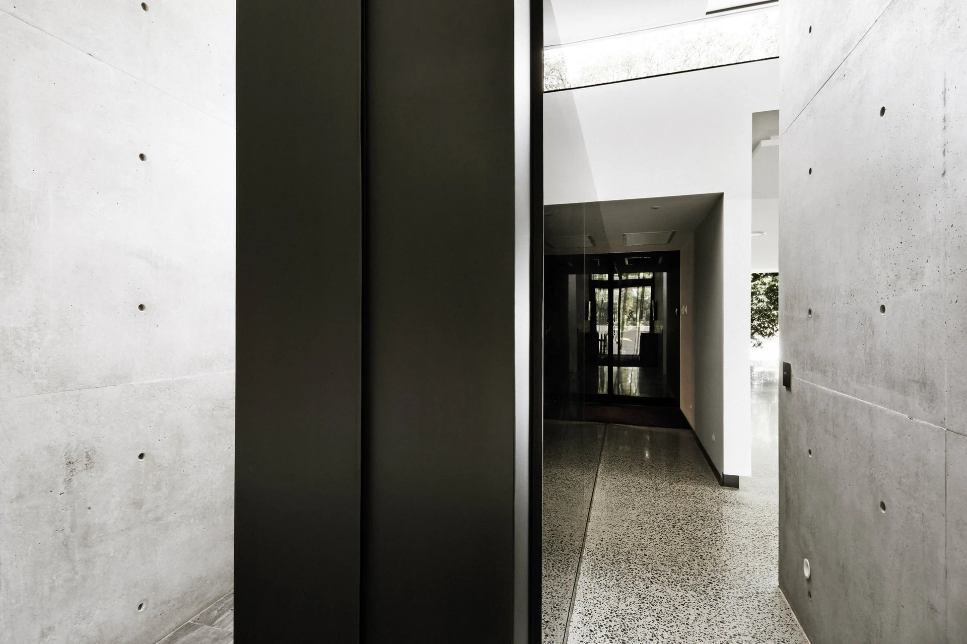 A minimalist modern entryway features matte grey concrete walls with tie holes and a polished terrazzo floor leading to a glass partition.