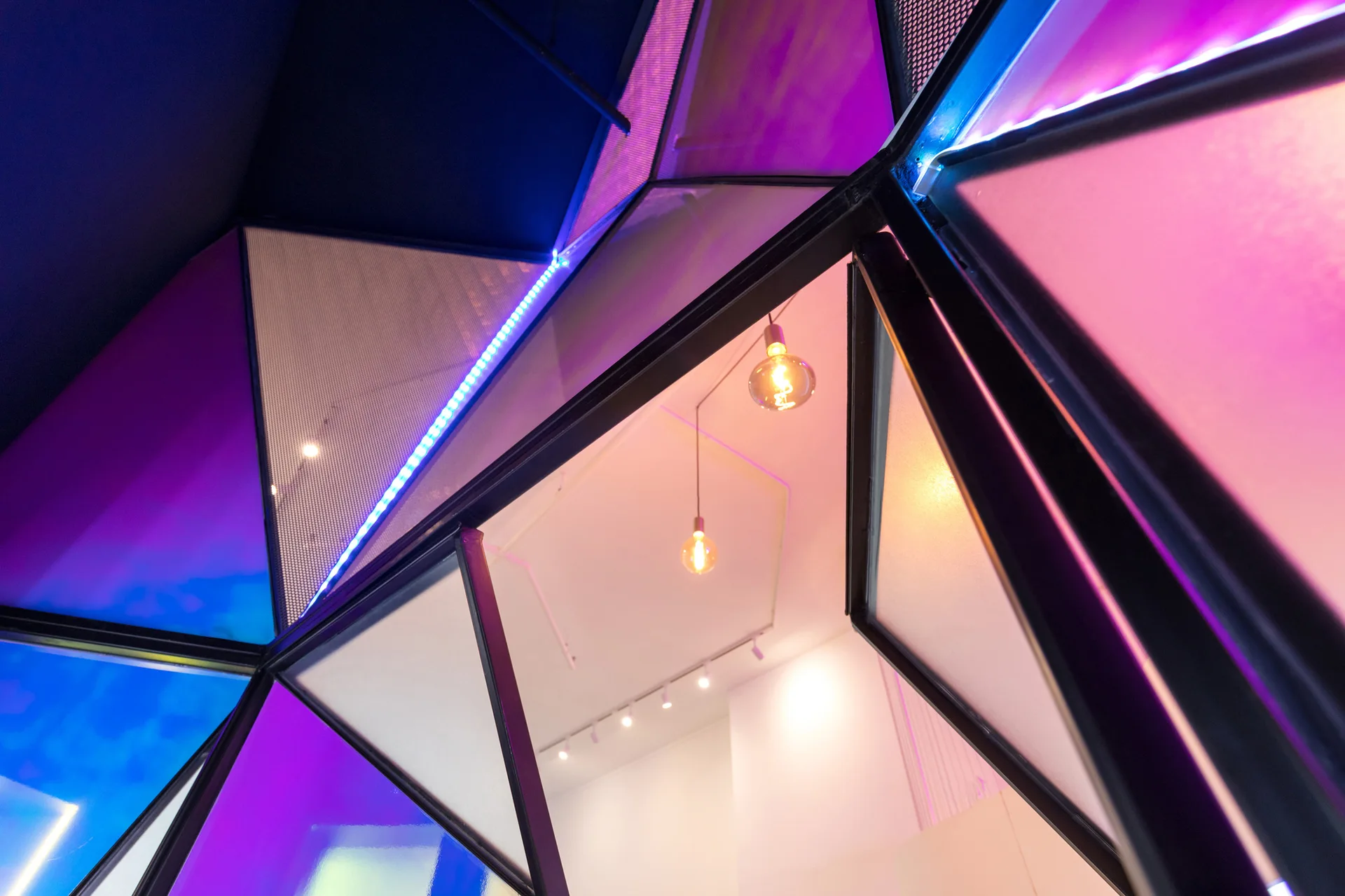 A geometric black-framed partition with triangular glass and mesh panels illuminated by purple and blue LED lights reveals simple pendant lights inside.