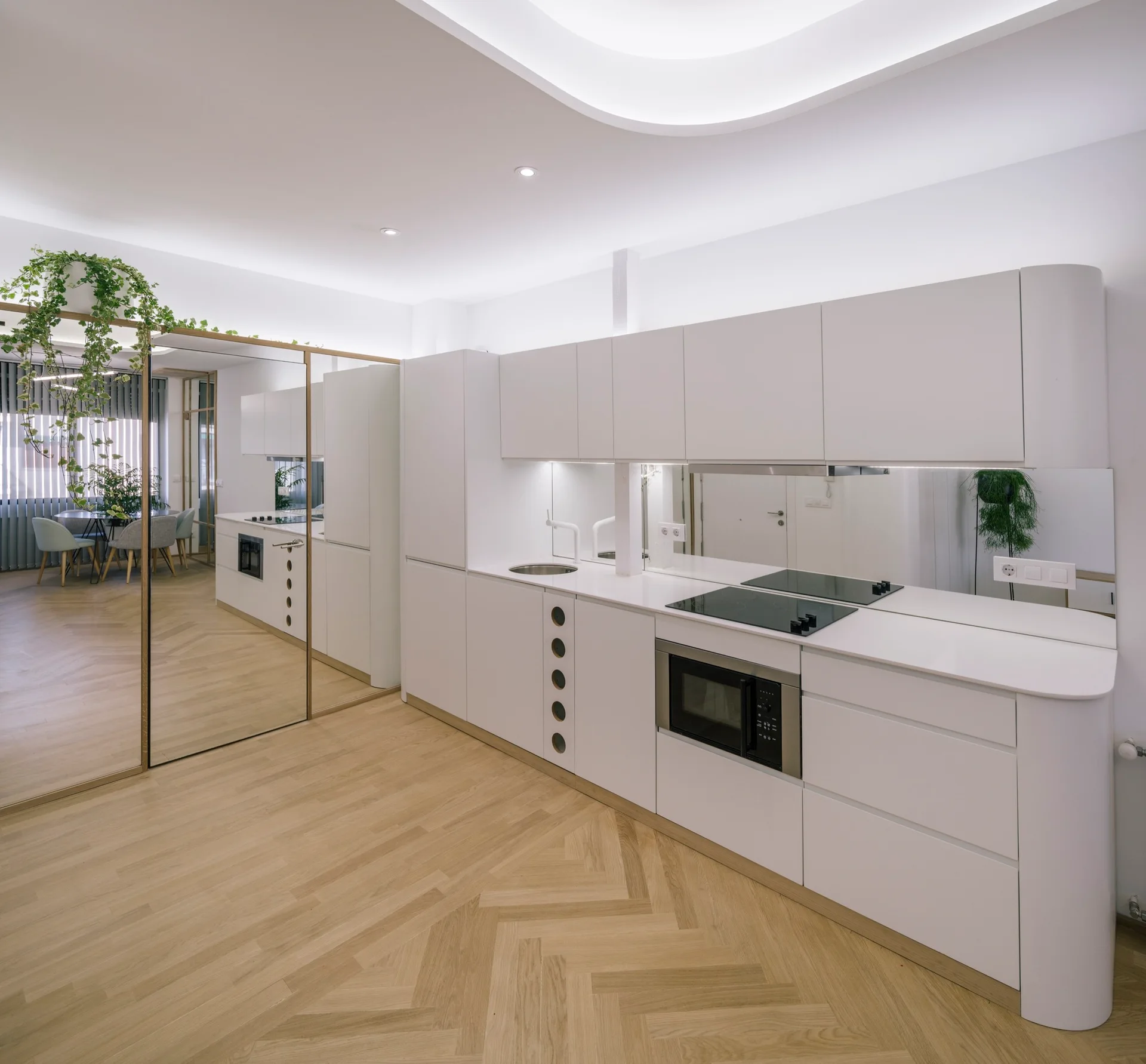 A minimalist white kitchen features handleless cabinets, a mirrored backsplash, and a light oak herringbone wood floor.