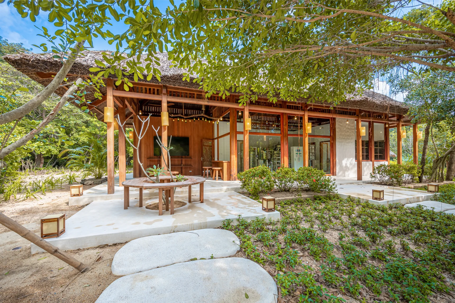 A tropical wooden pavilion features a thatched roof, hanging lanterns, and a circular bench wrapping around a small tree on a paved patio.
