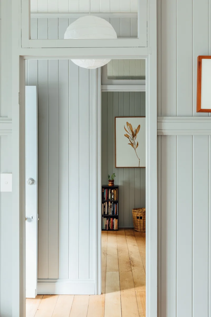 A view through a doorway shows light grey vertical wood paneling, natural wood floors, a paper globe pendant light, and a framed botanical art print.