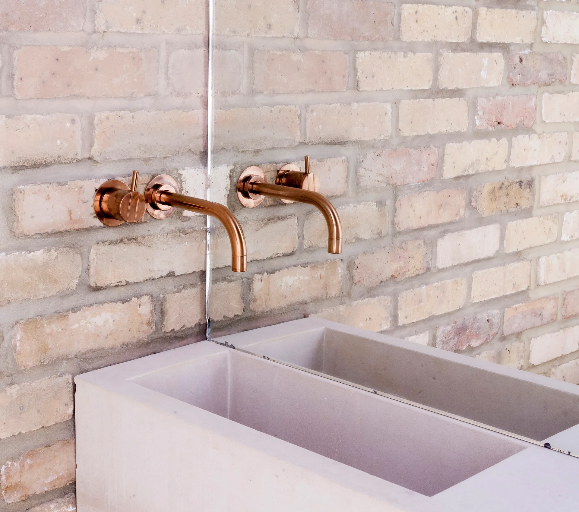 A cast concrete trough sink sits beneath wall-mounted copper faucets on an exposed tan brick wall bisected by a mirror.