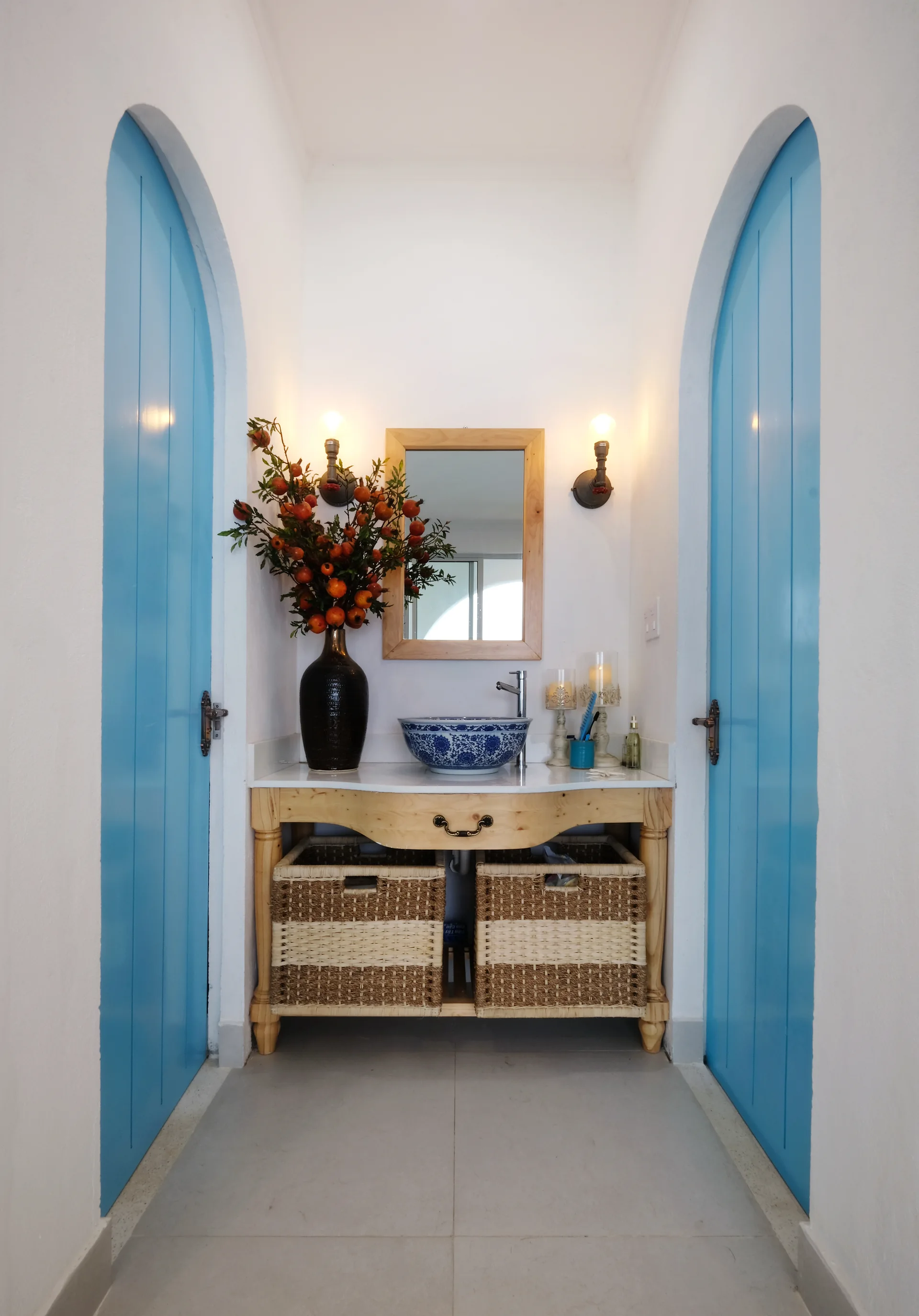 A wooden vanity with a patterned blue vessel sink is flanked by two vibrant blue arched doors in a symmetrical white-walled powder room.
