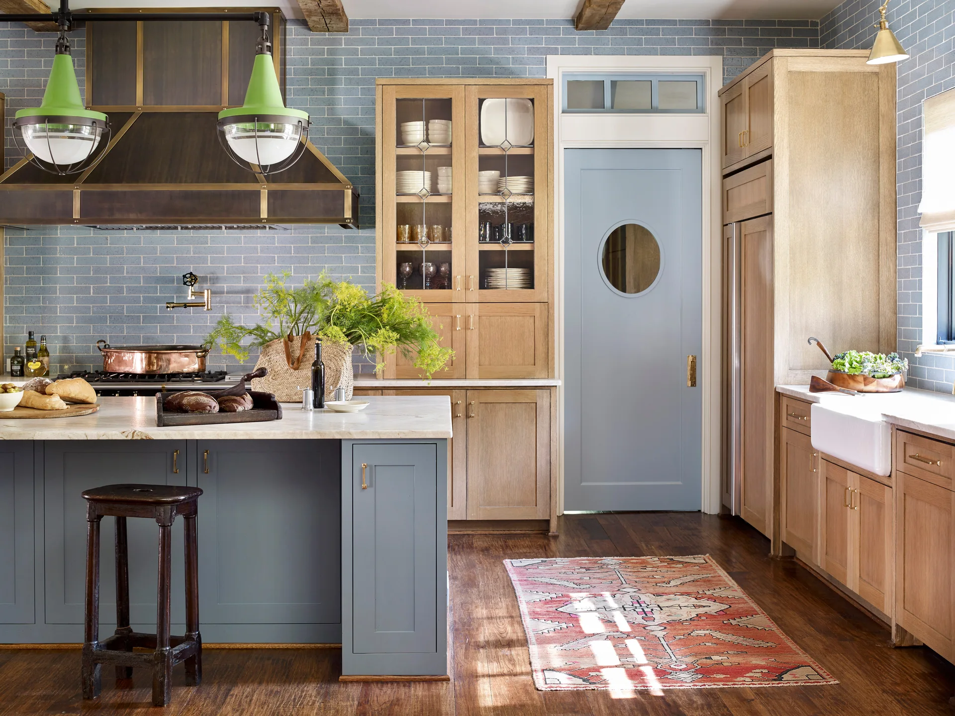 A residential kitchen features natural wood cabinets, a blue kitchen island, a matching blue tiled wall, and vibrant green pendant lights.