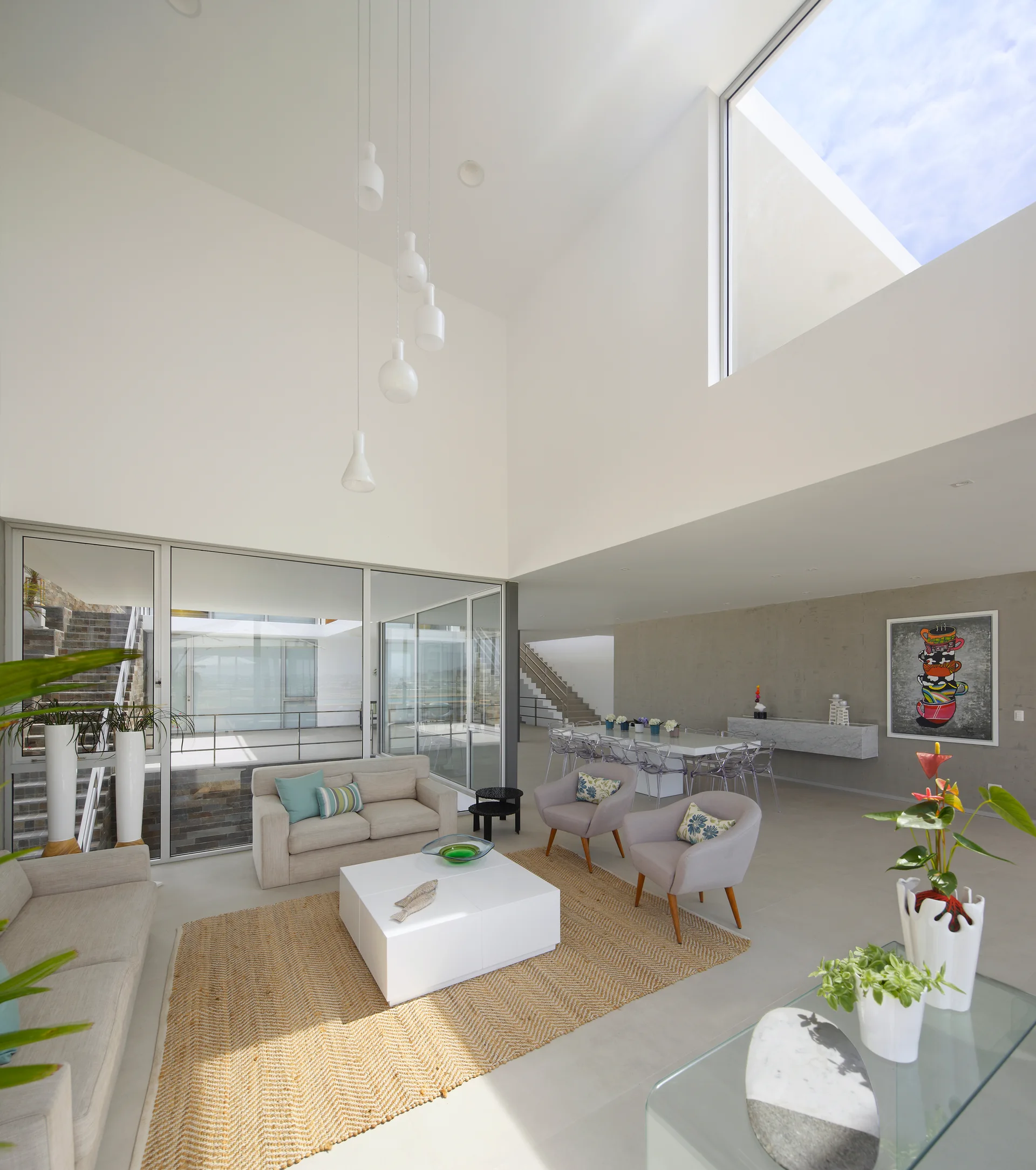 A minimalist living room features a double-height ceiling with a large skylight, a beige sofa on a chevron woven rug, and a dining area.