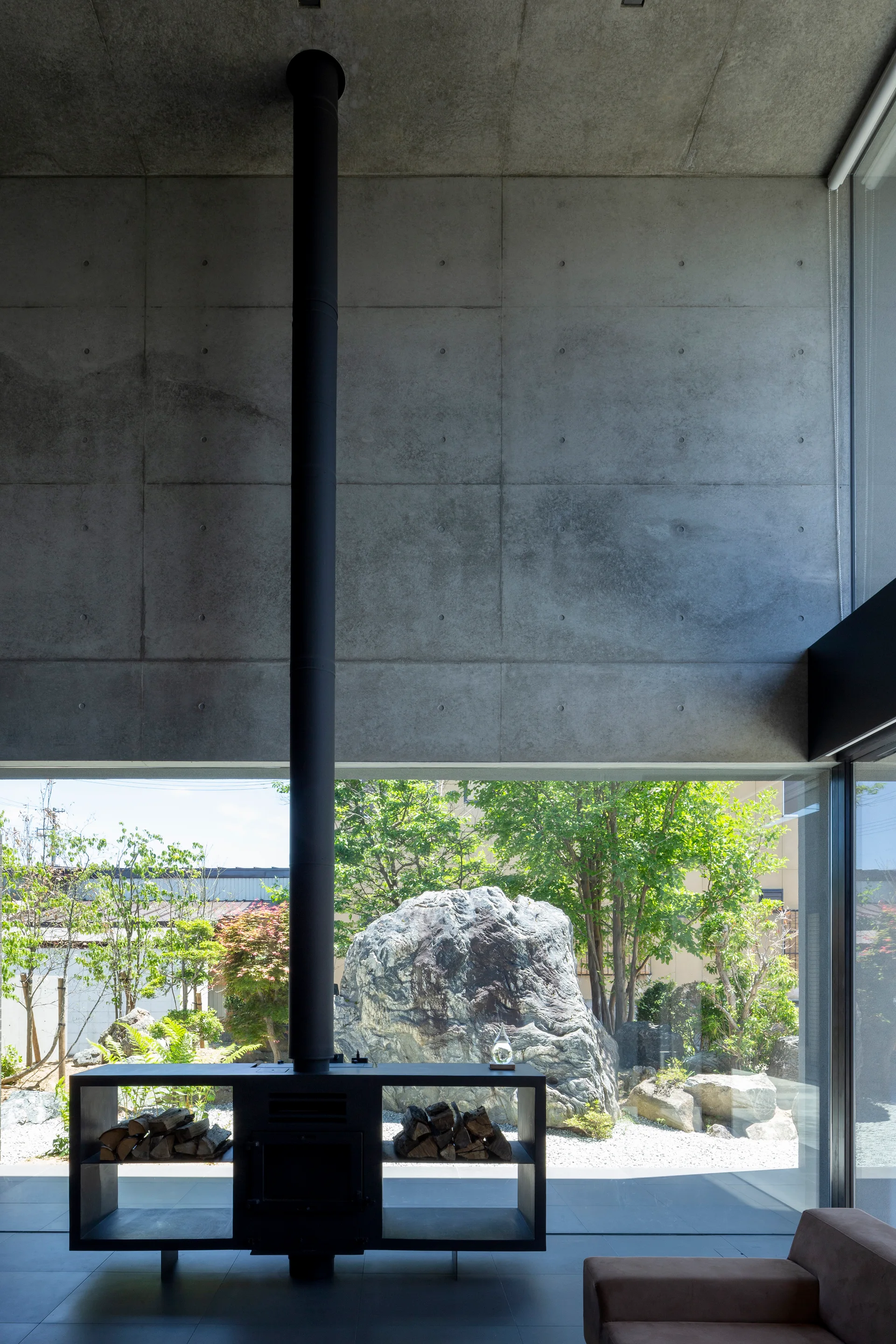 A minimalist living room features a raw concrete wall, a black metal fireplace with integrated wood storage, and glass windows overlooking a stone garden.
