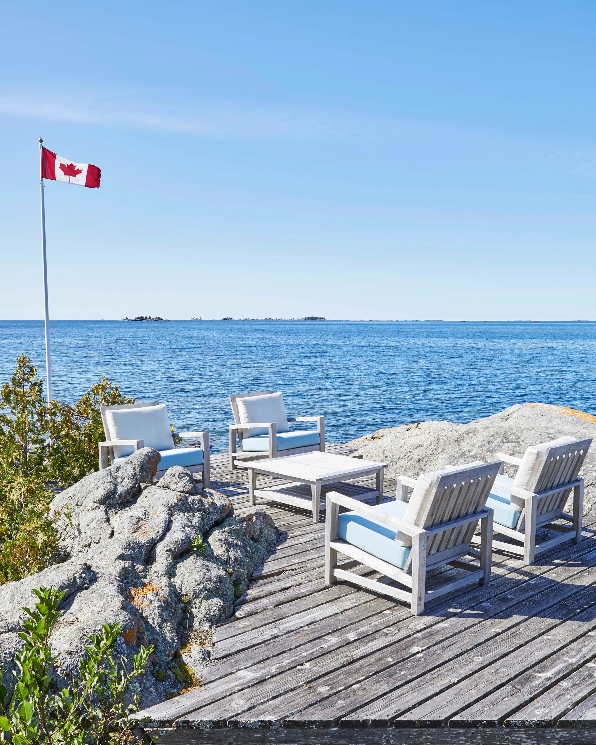 Four weathered wooden lounge chairs with light cushions surround a low table on a wood deck next to natural rock and calm blue water.