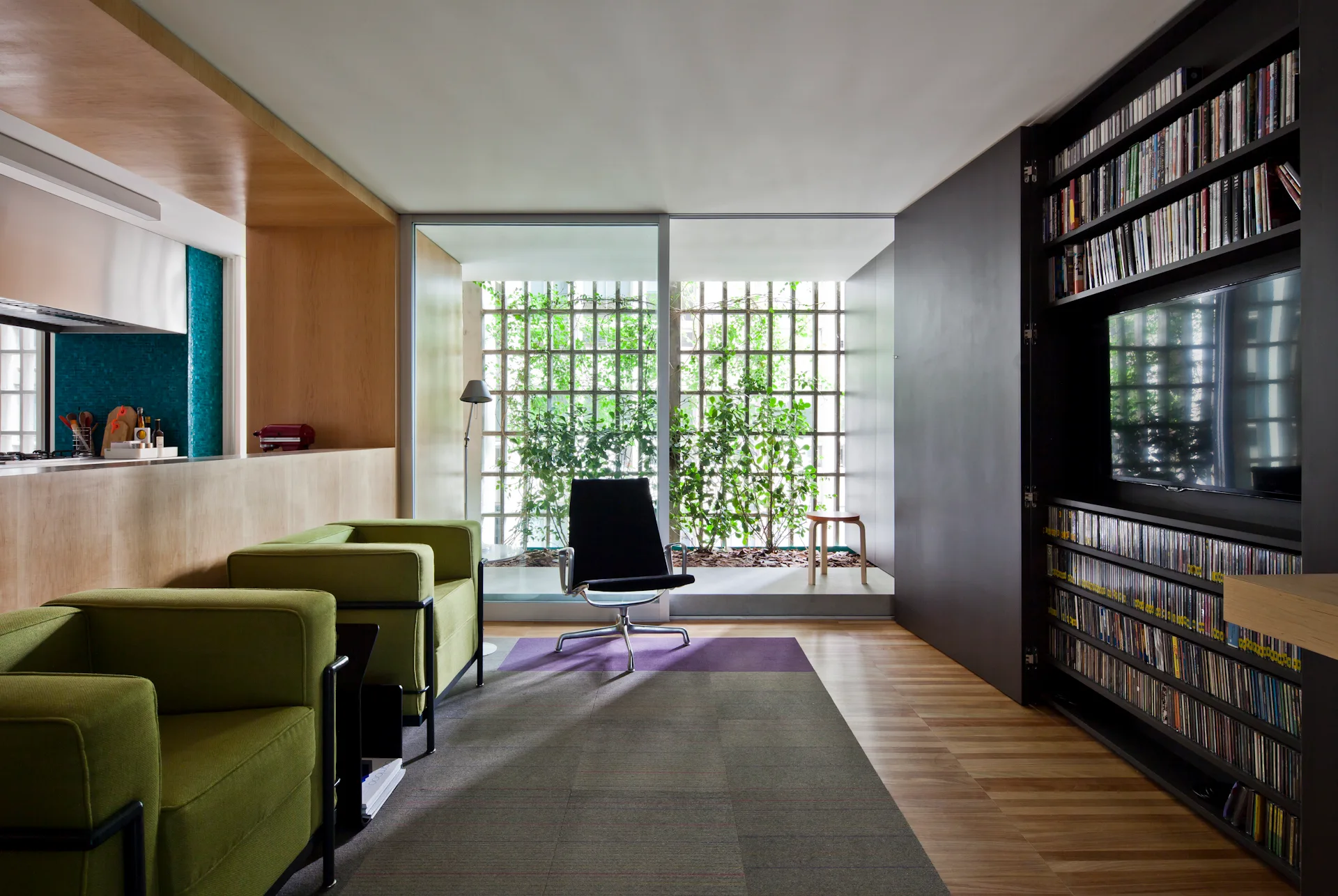 A modern living room features olive green armchairs, a large dark grey built-in media cabinet with shelving, and a glass wall looking out to a garden.