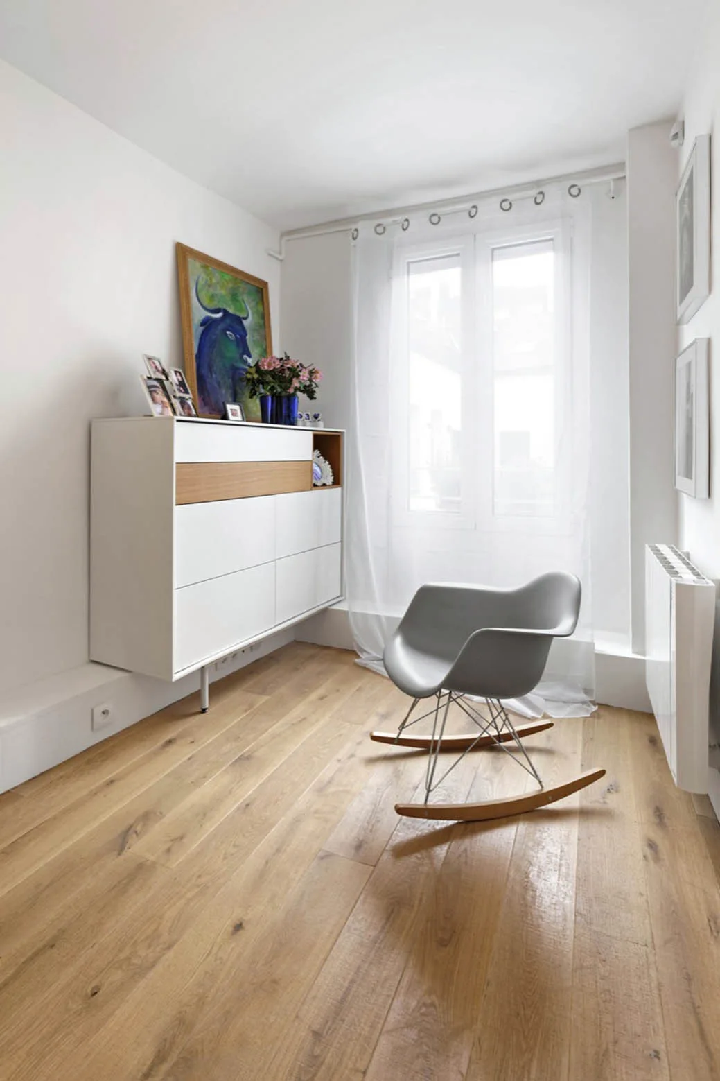 A minimalist room features a grey molded rocking chair on light wood floors near a white wall-mounted storage cabinet and a sunlit window.