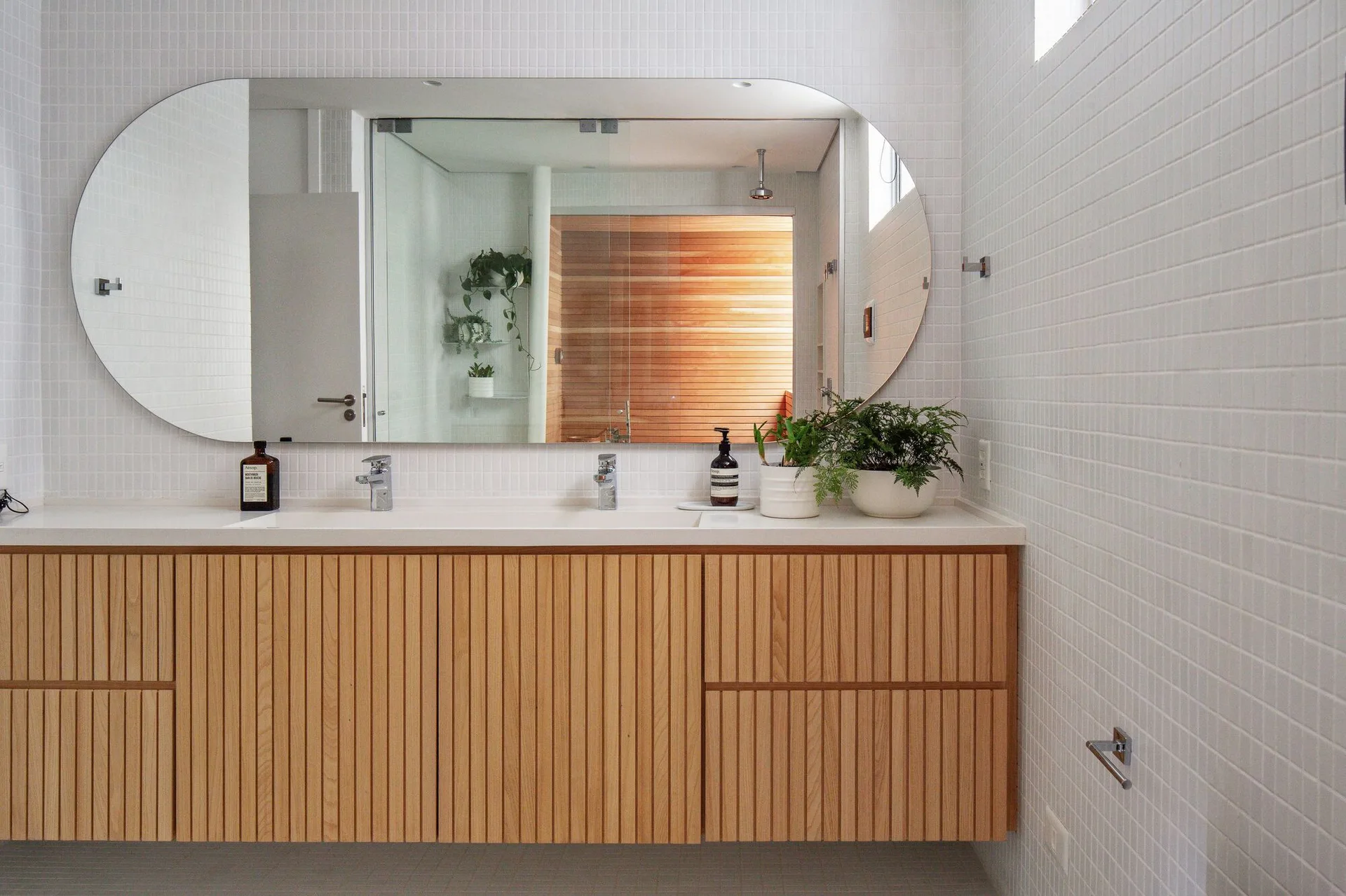 A floating slatted-wood vanity sits beneath a large pill-shaped mirror on a wall covered in small white square tiles.