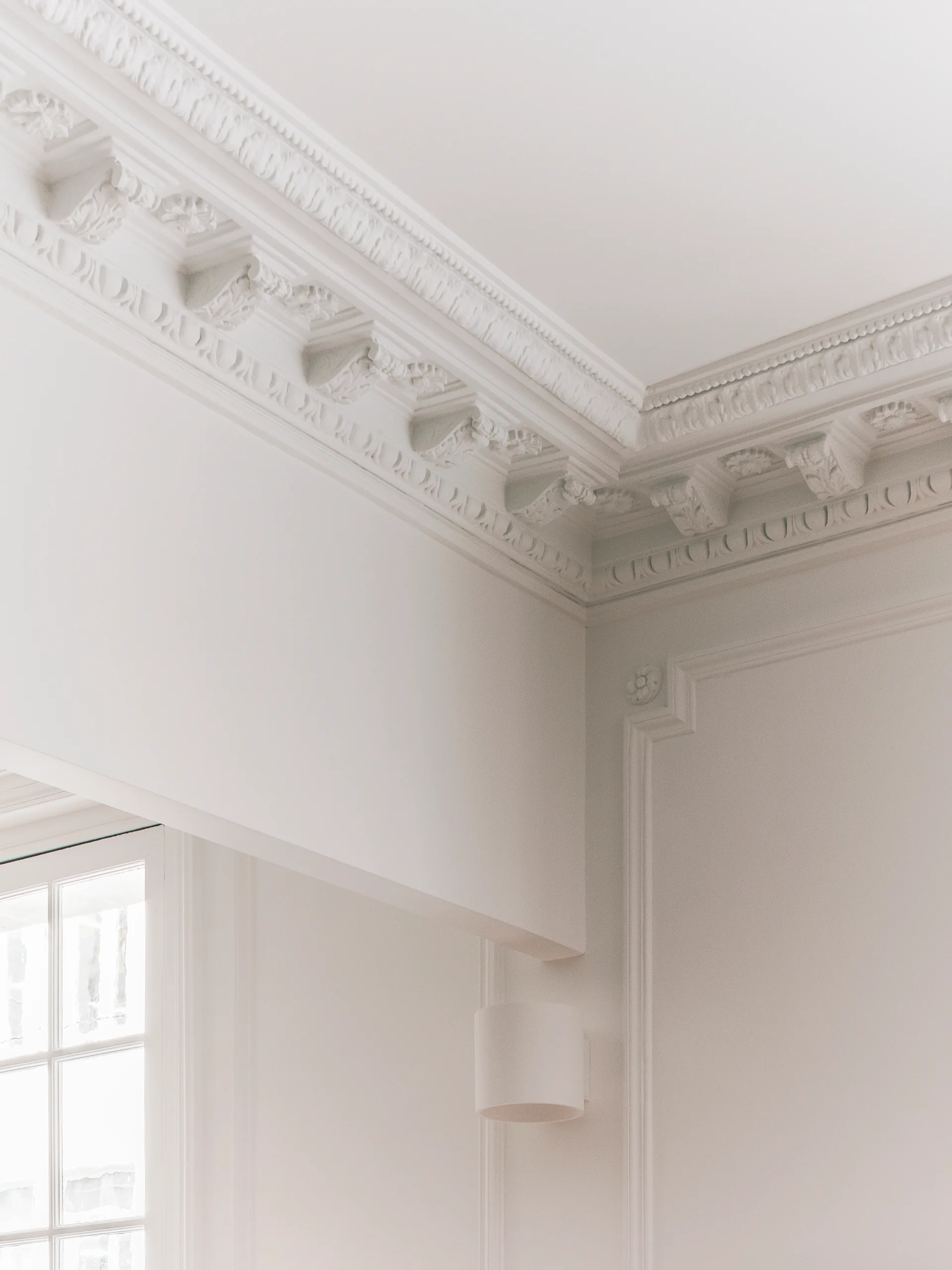 Architectural detail of ornate white plaster crown molding, traditional wall panels, a modern white cylindrical sconce, and a window frame.
