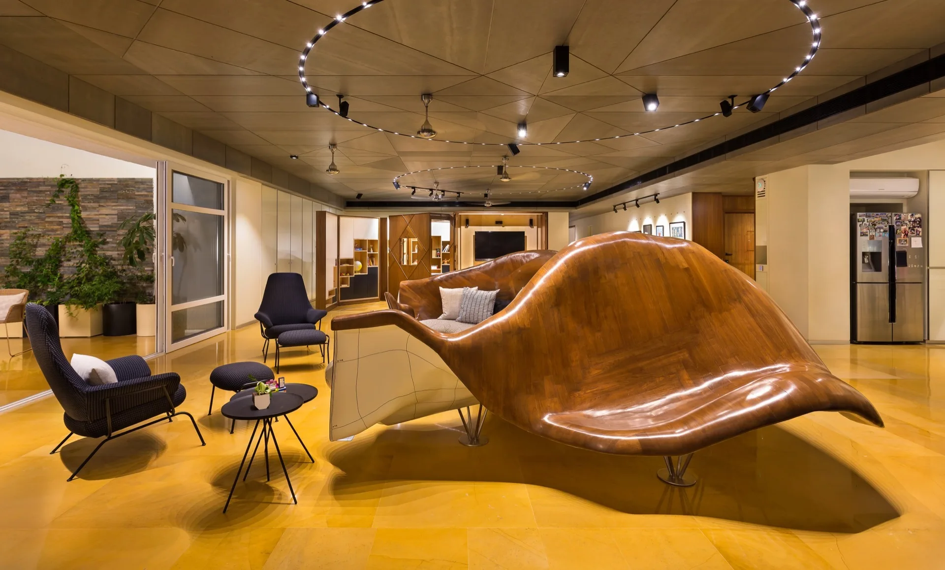 A spacious living room features a large curved wood sculptural sofa on yellow stone tile flooring beneath a geometric paneled ceiling.