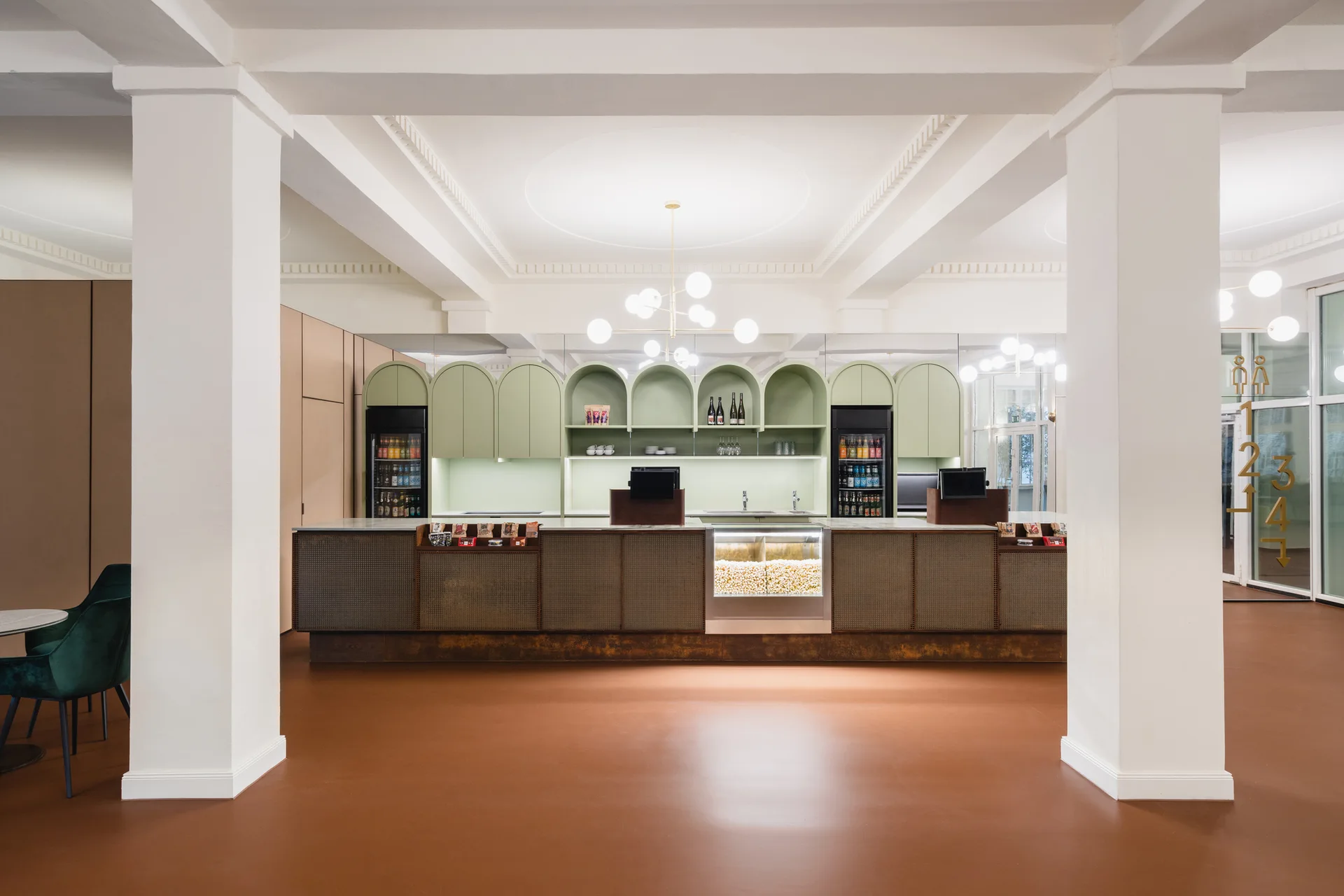 A modern commercial bar area features a long mesh counter, sage green arched shelving built into the wall, large white columns, and rust-colored flooring.