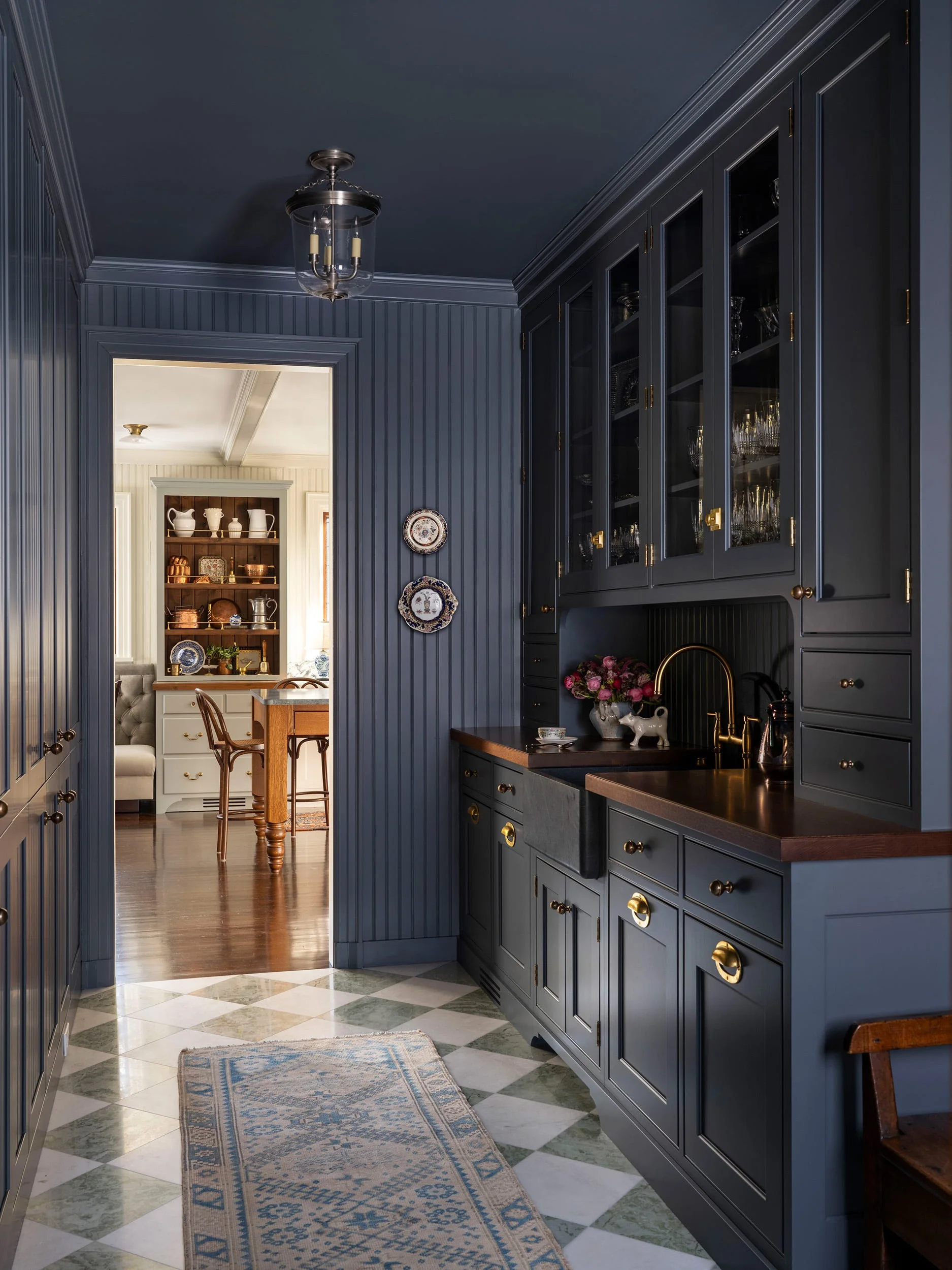A dark blue butler's pantry features matching painted beadboard walls, wood countertops, a farmhouse sink, and a green and white checkerboard stone floor.