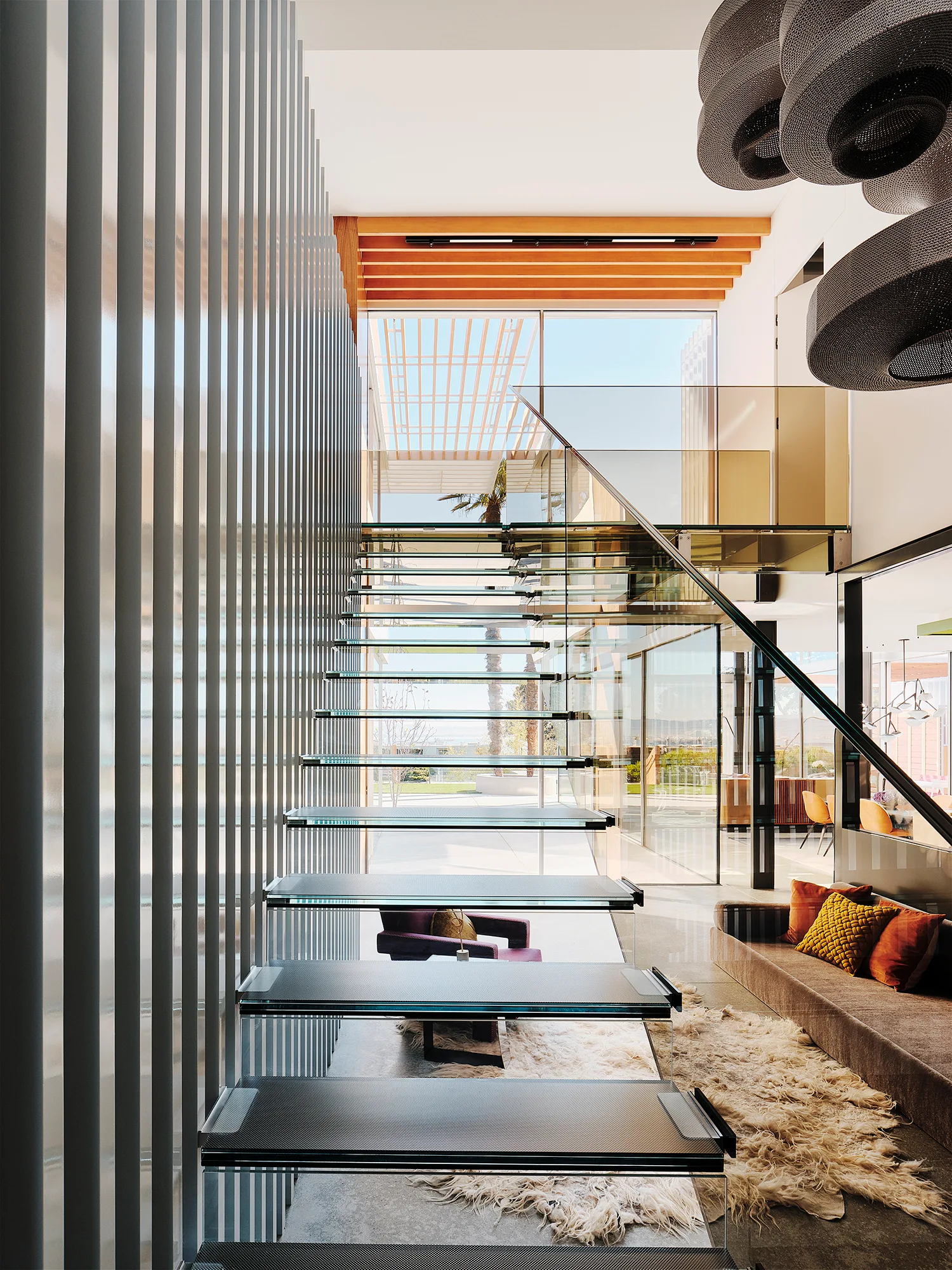 A modern foyer featuring a floating glass staircase next to a vertical slatted wall, with a low sofa and shag rug underneath.