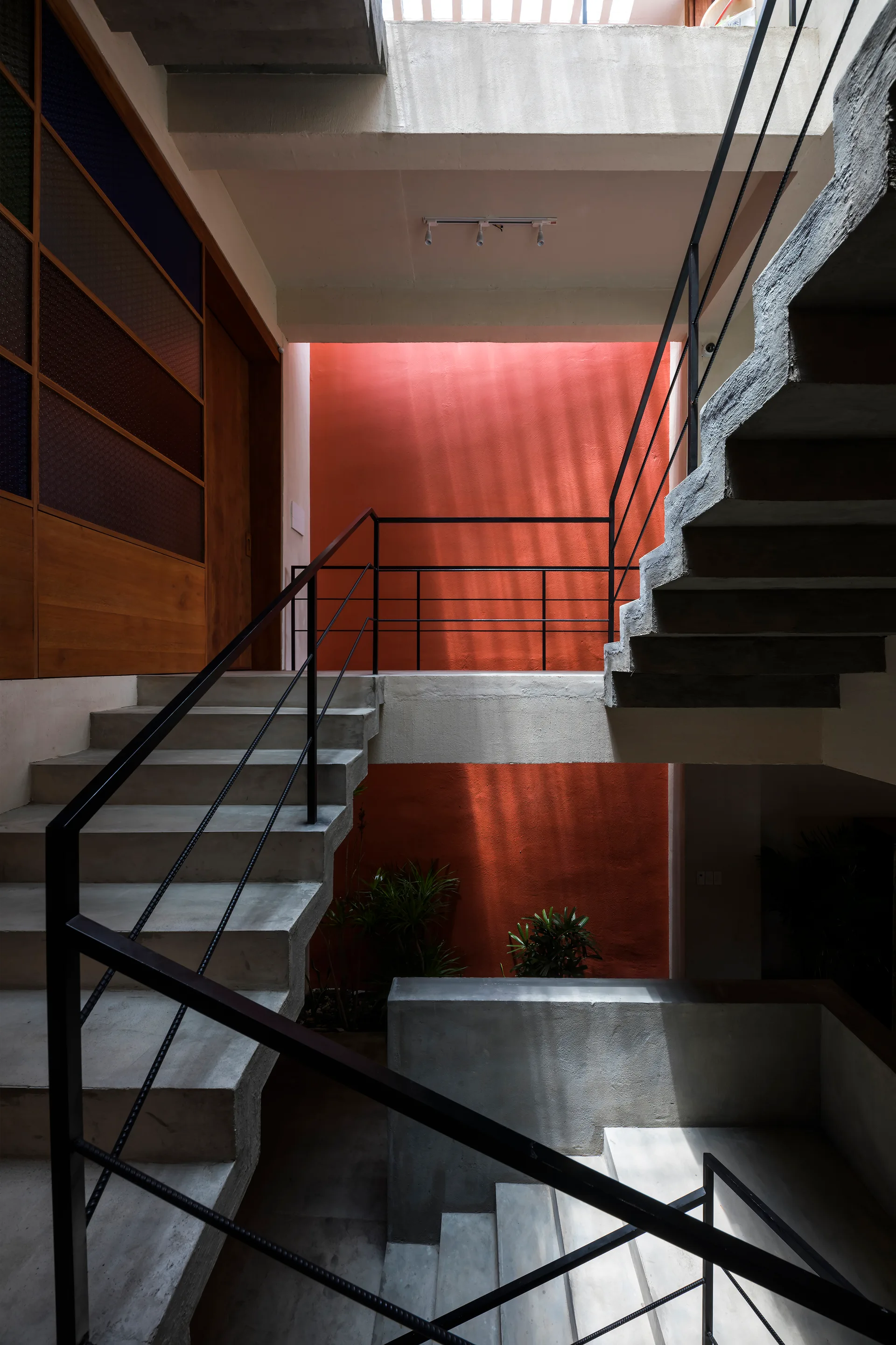 A multi-level concrete staircase with black metal railings stands in an open atrium opposite a tall, matte terracotta-orange painted accent wall.
