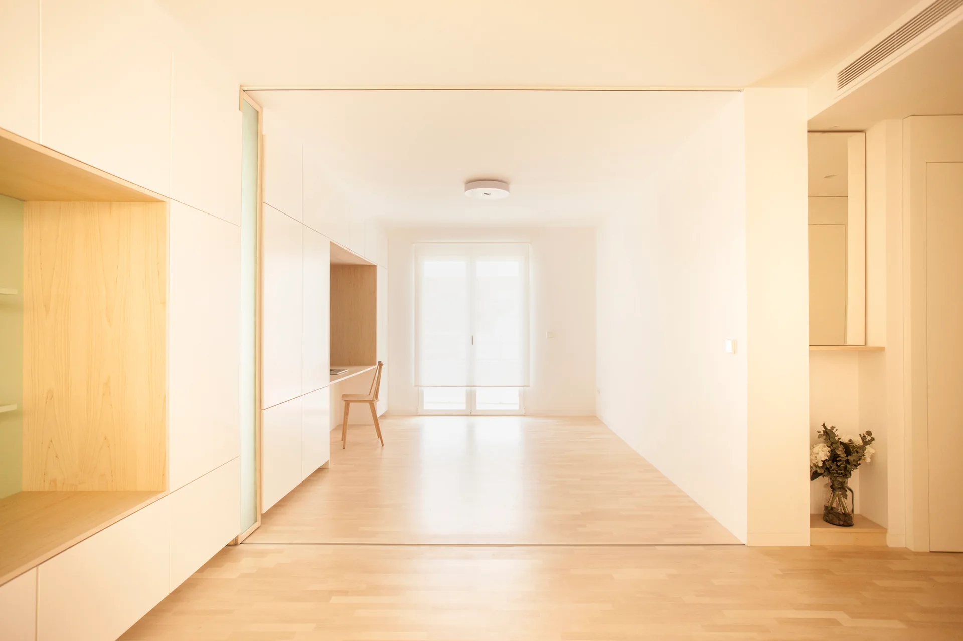 Minimalist room featuring matte white built-in cabinets with integrated light wood desk niches and a wooden chair on blonde wood floors.