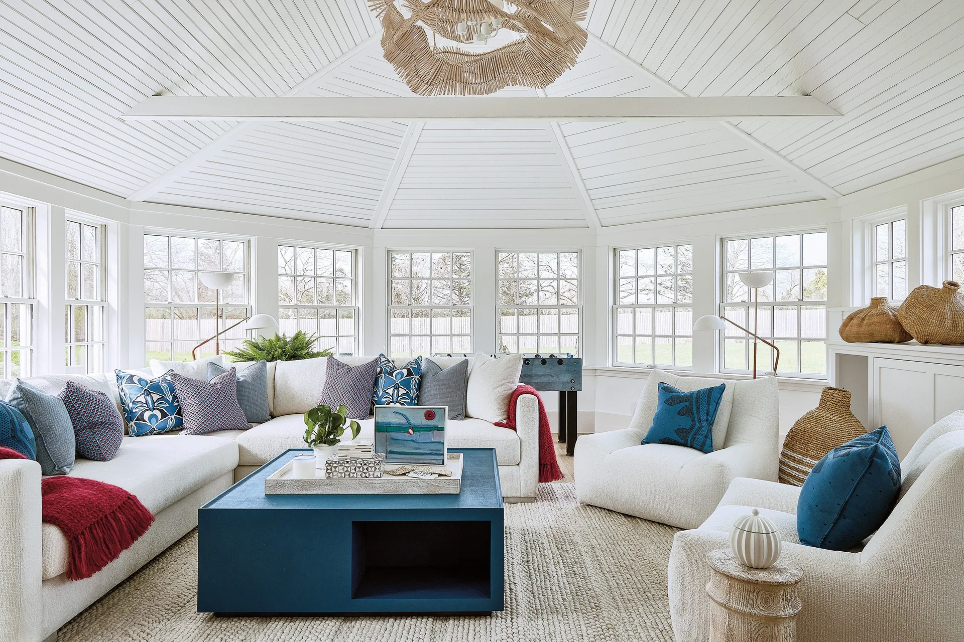 A sunroom with a vaulted white plank ceiling, a cream sectional sofa, a bold blue coffee table, and multiple windows.