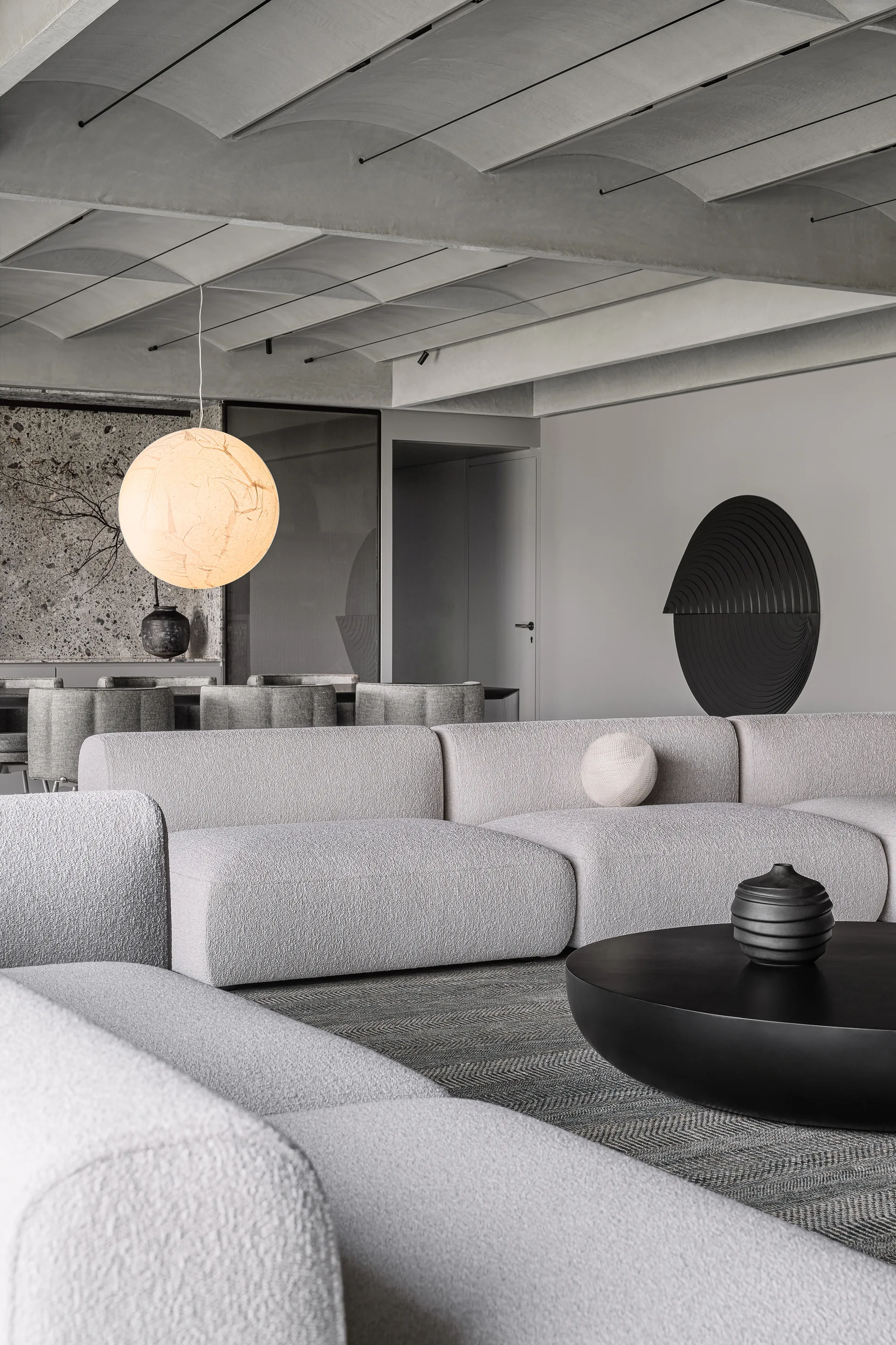 A minimalist living room featuring a large cream boucle sectional sofa, a round black coffee table, and a segmented vaulted concrete ceiling.