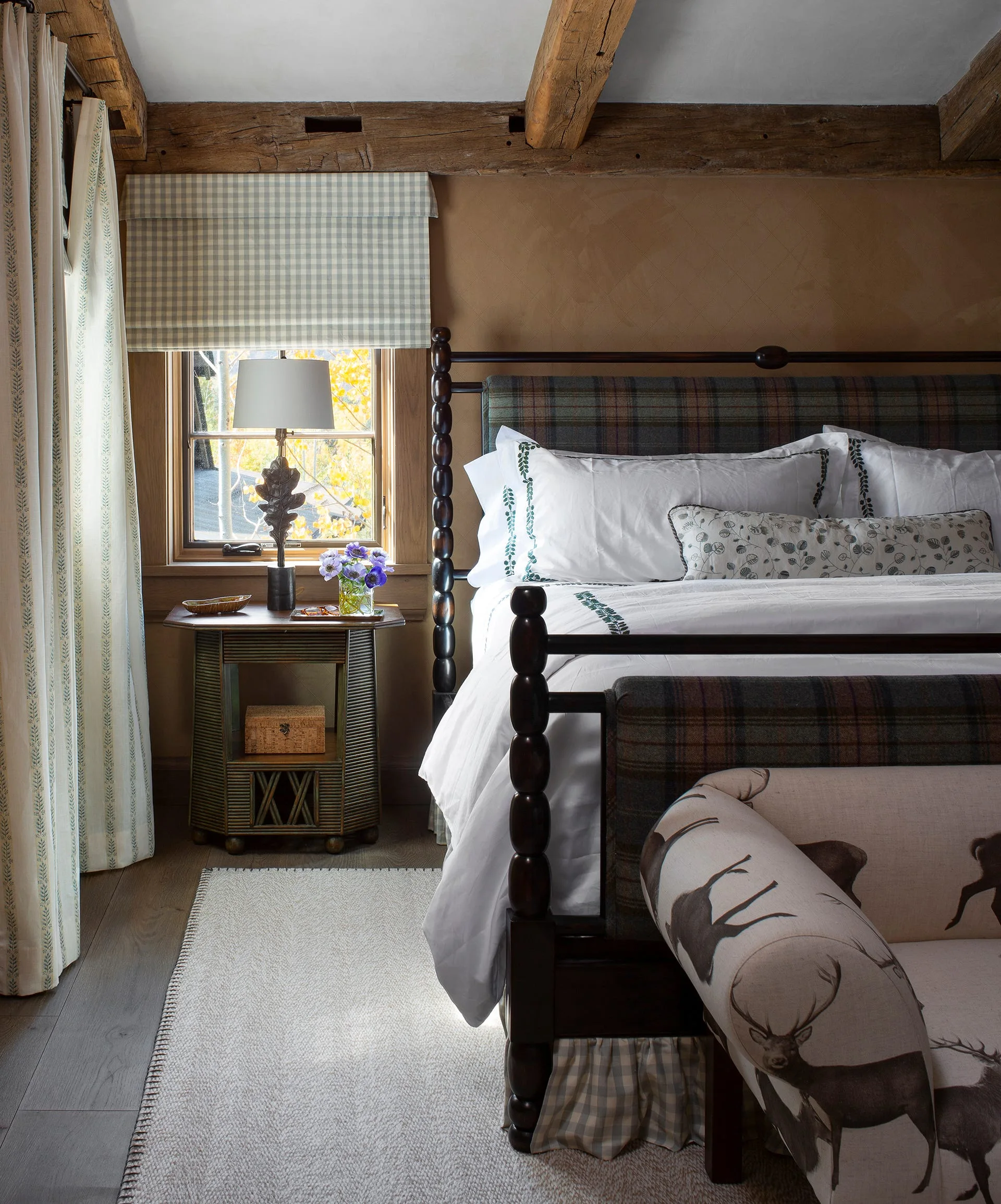 A rustic bedroom features a dark wood spindle bed with a plaid headboard, a deer-print bench, and a small nightstand next to a window.