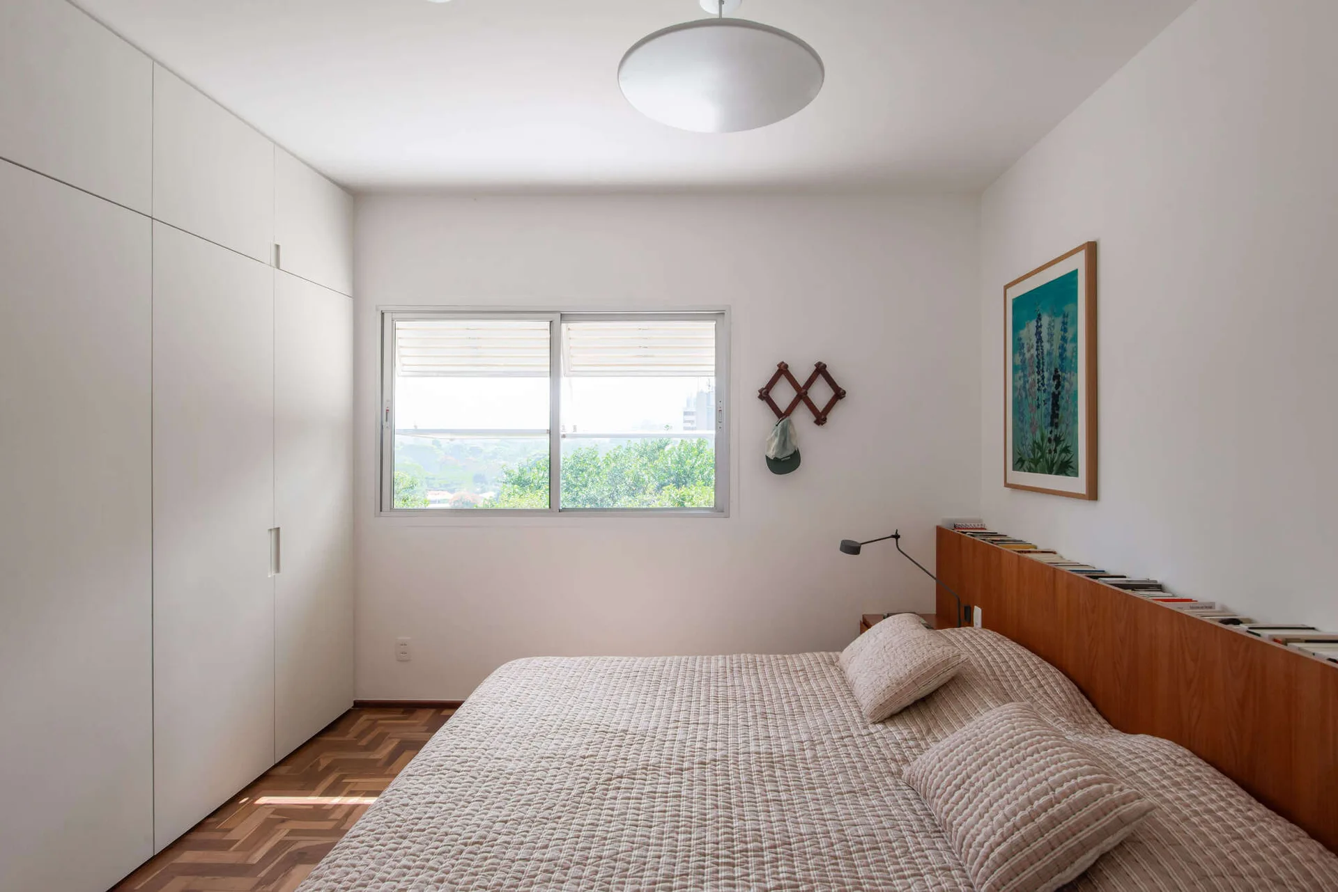 A minimalist bedroom features a bed with a quilted beige coverlet, a wood-toned headboard storing books, a built-in wardrobe, and herringbone flooring.