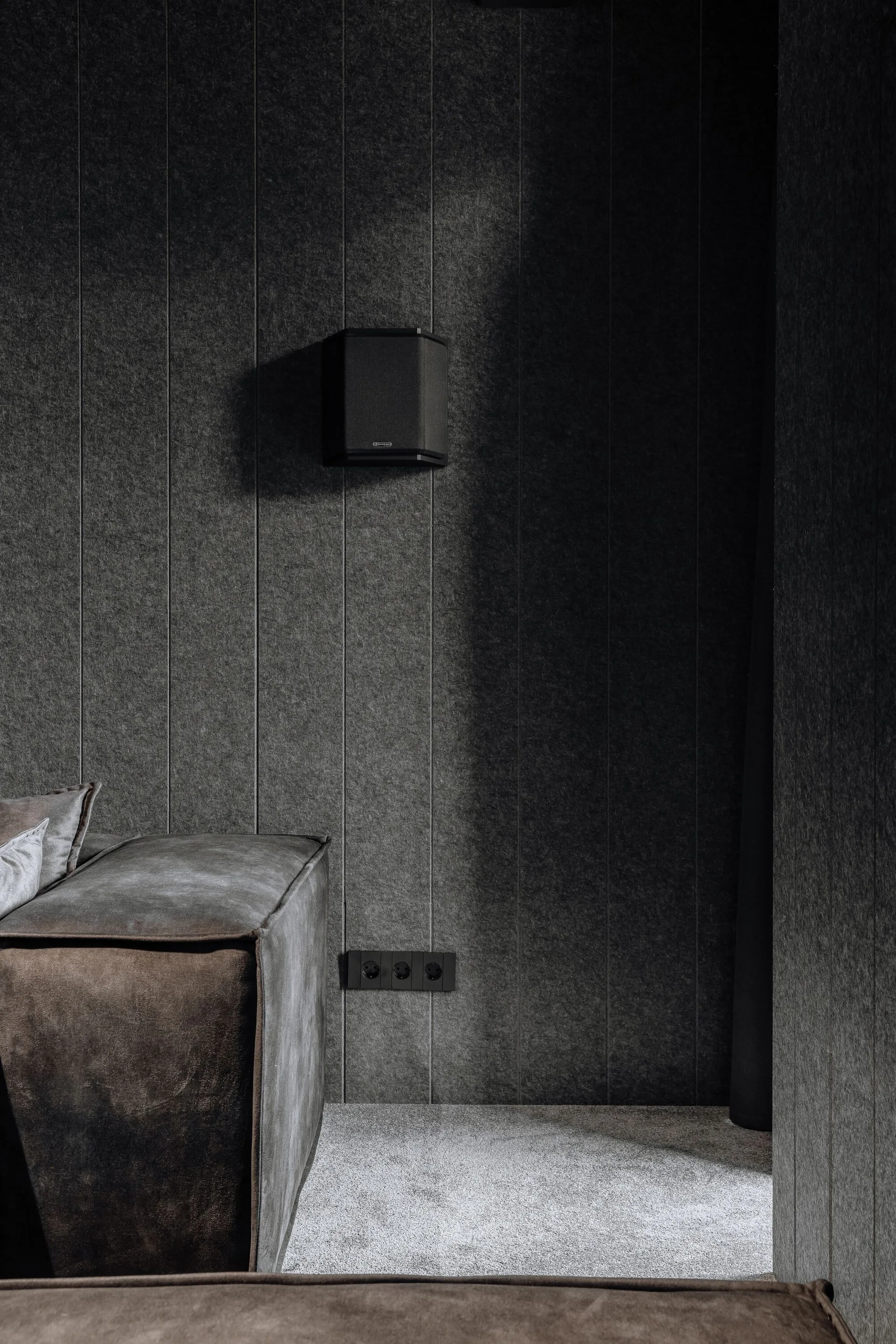 A close-up of a dark room featuring vertical charcoal felt wall panels, a black wall speaker, and a gray suede sofa corner.