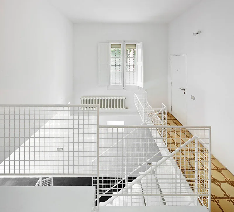 A minimalist white staircase and landing featuring a white wire grid railing, a shuttered window, and patterned geometric floor tiles.