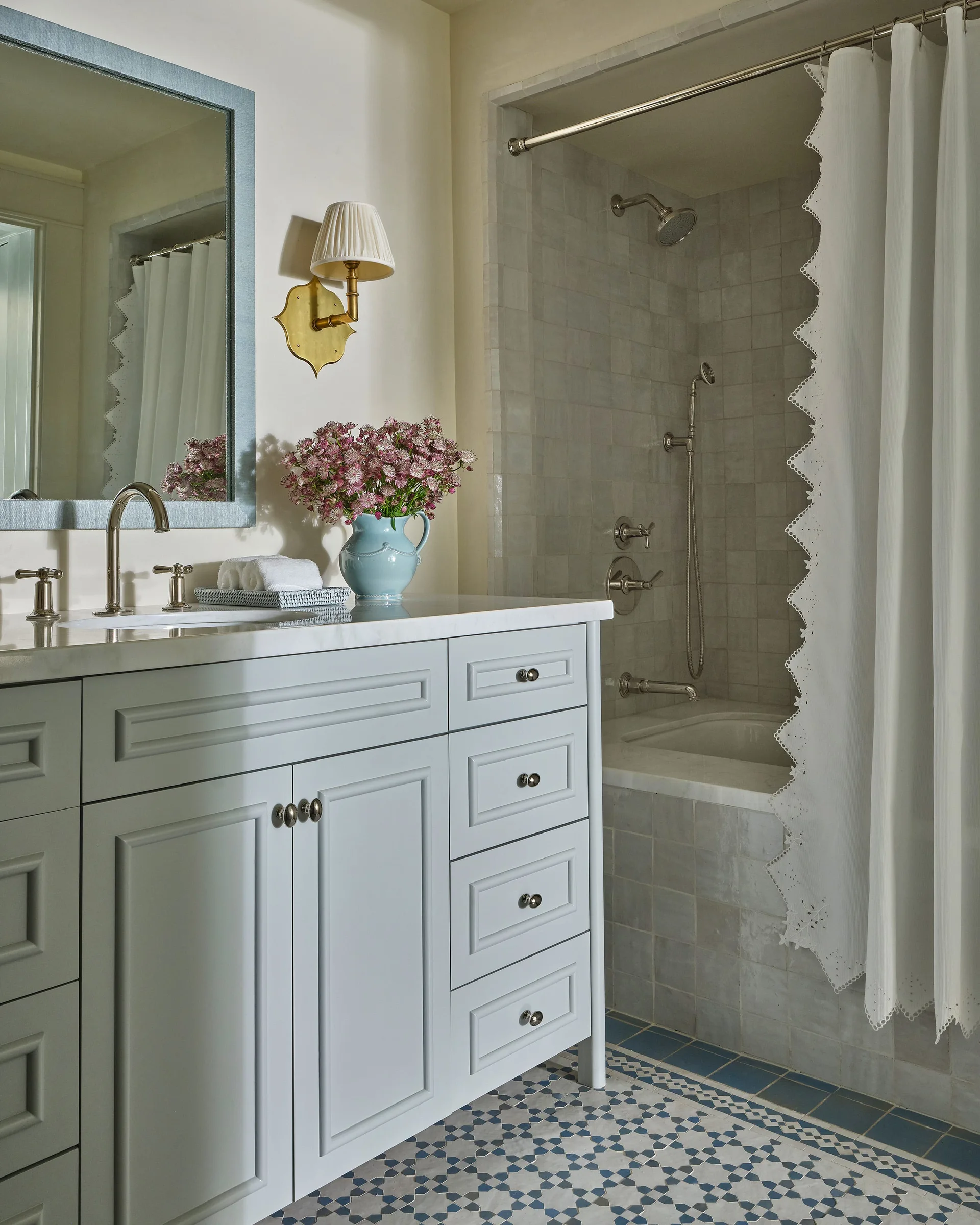A bathroom with a light blue vanity, patterned floor tile, and a tiled shower enclosed by a white scalloped shower curtain.