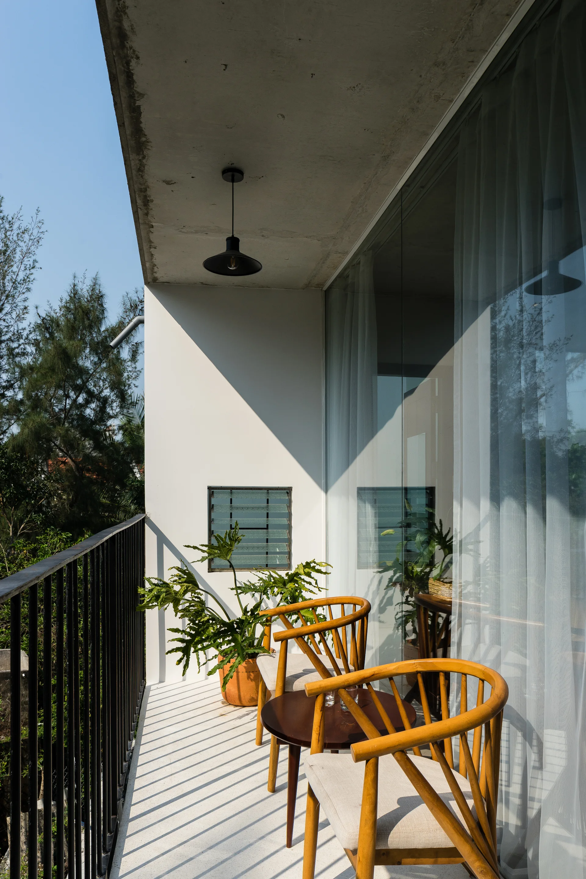 Two wooden armchairs and a small round table sit on a white concrete balcony beneath an exposed concrete ceiling and black pendant light.
