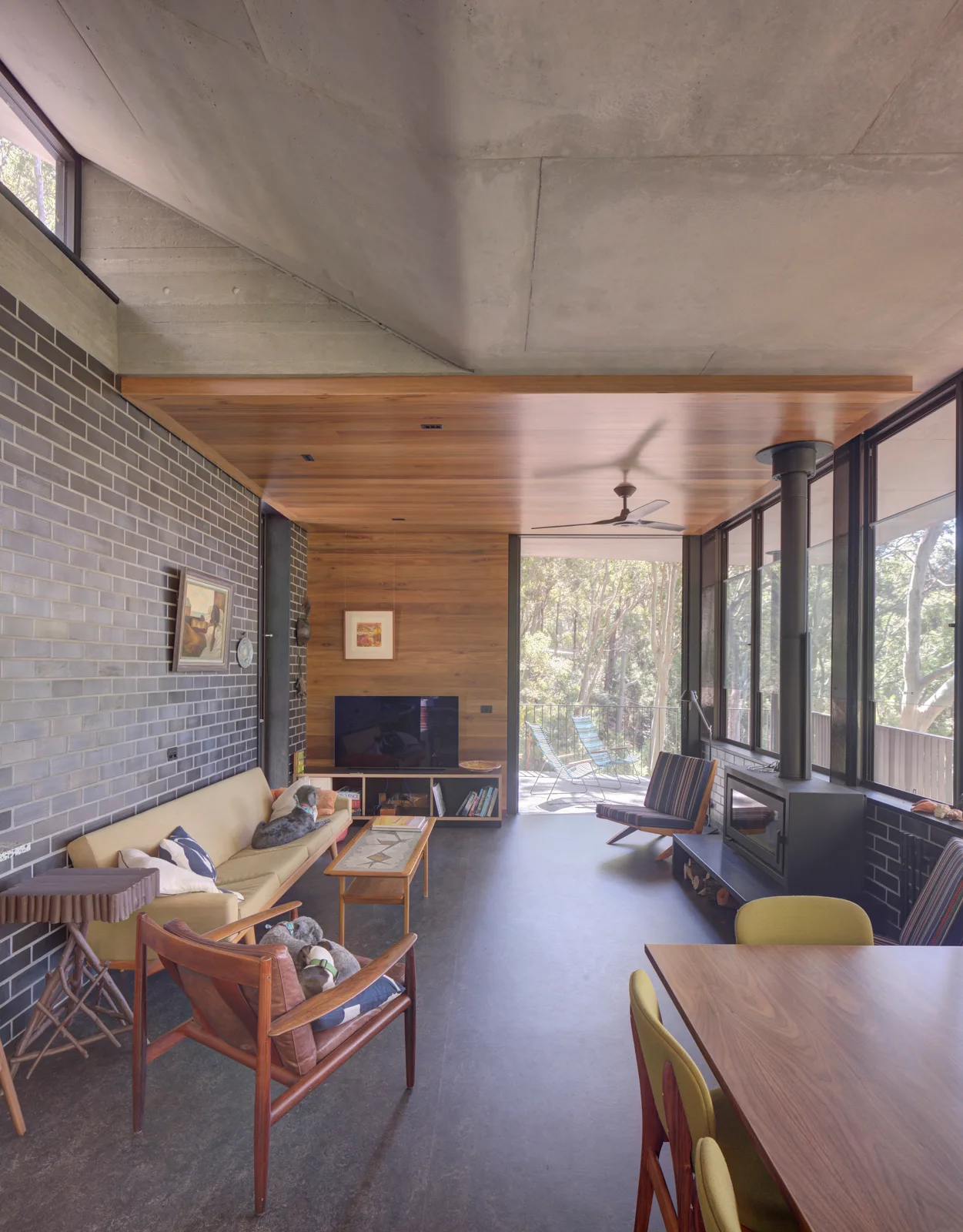 A midcentury modern living room features a dark grey brick wall, a raw concrete ceiling, a wood-paneled soffit, and a yellow sofa.