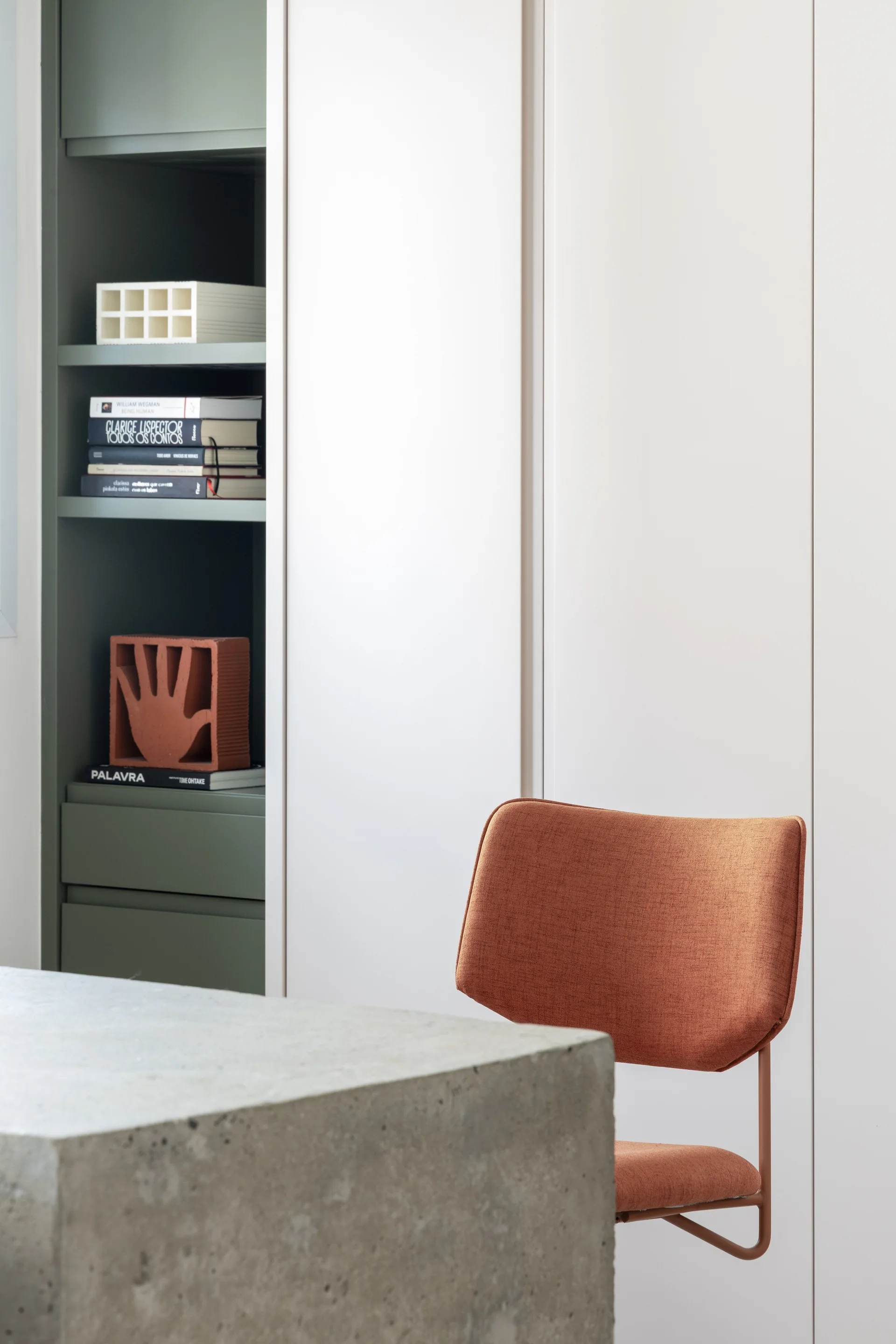 A minimalist study corner features a textured concrete desk, an orange fabric chair, and white built-in cabinets with a recessed sage green bookshelf.