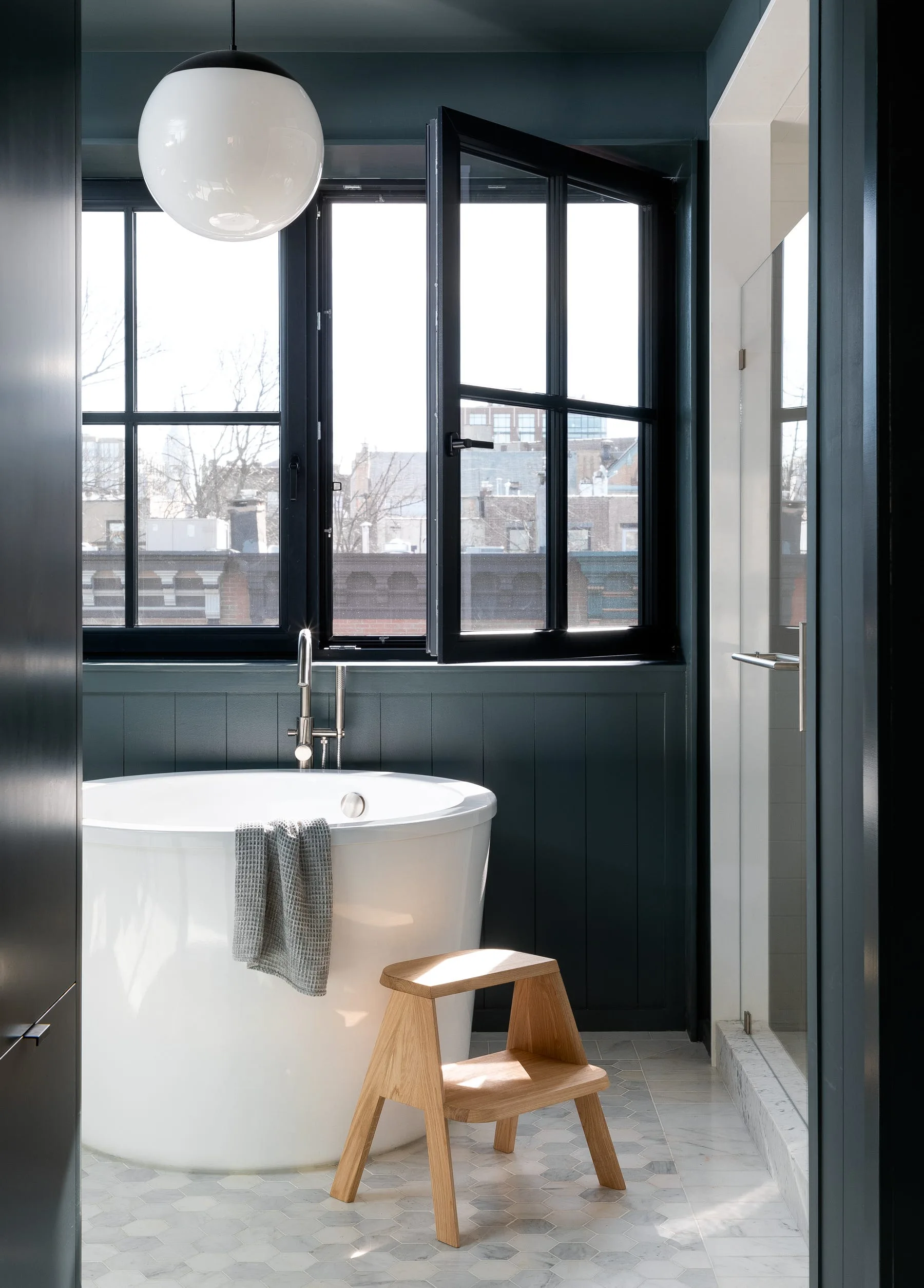 A round white soaking tub sits in a bathroom with dark teal vertical paneling, a wooden stool, and hexagonal marble floor tiles.