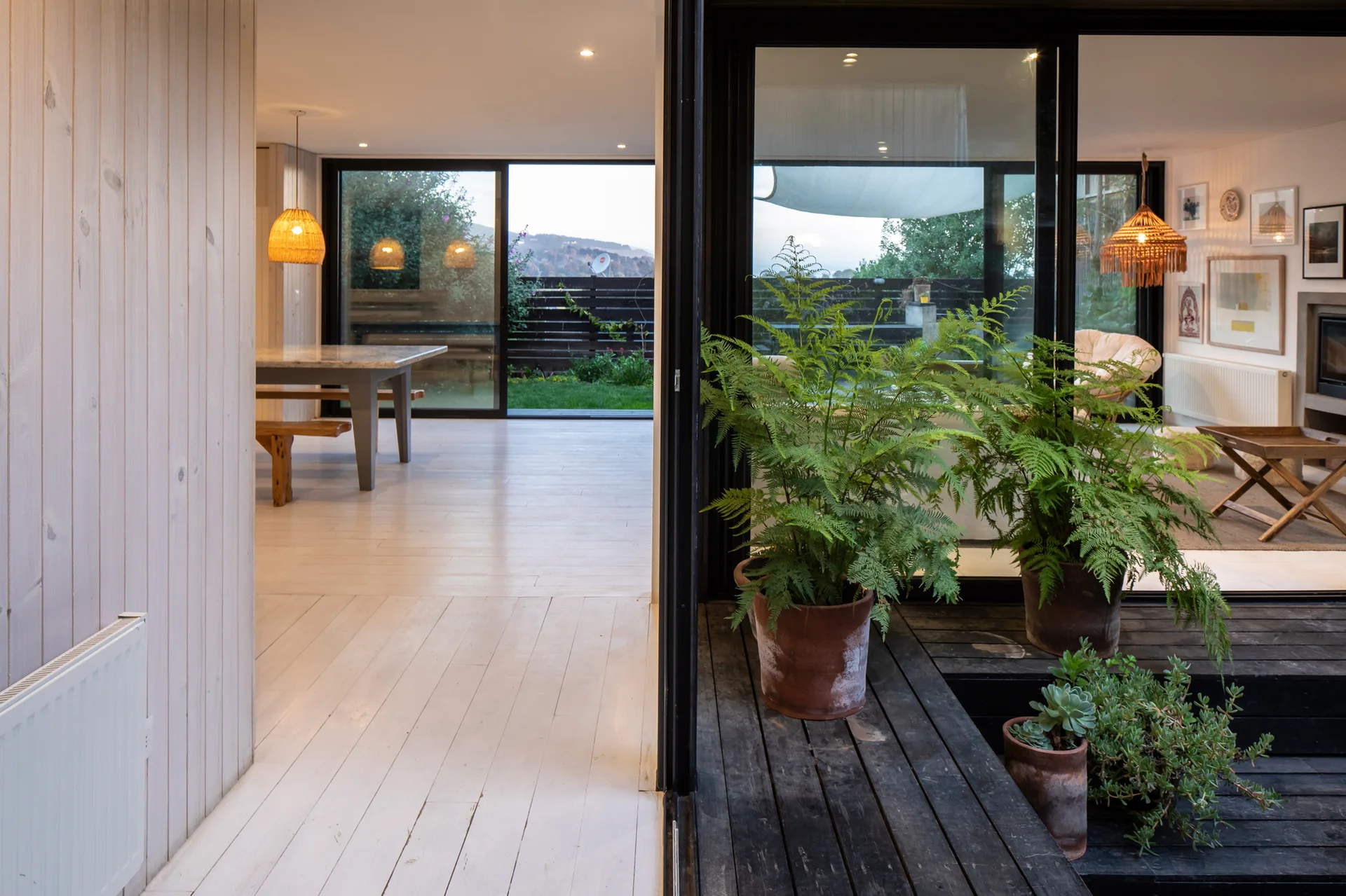 White-washed wood plank flooring and walls lead to black-framed sliding glass doors overlooking potted ferns on a dark wood step.