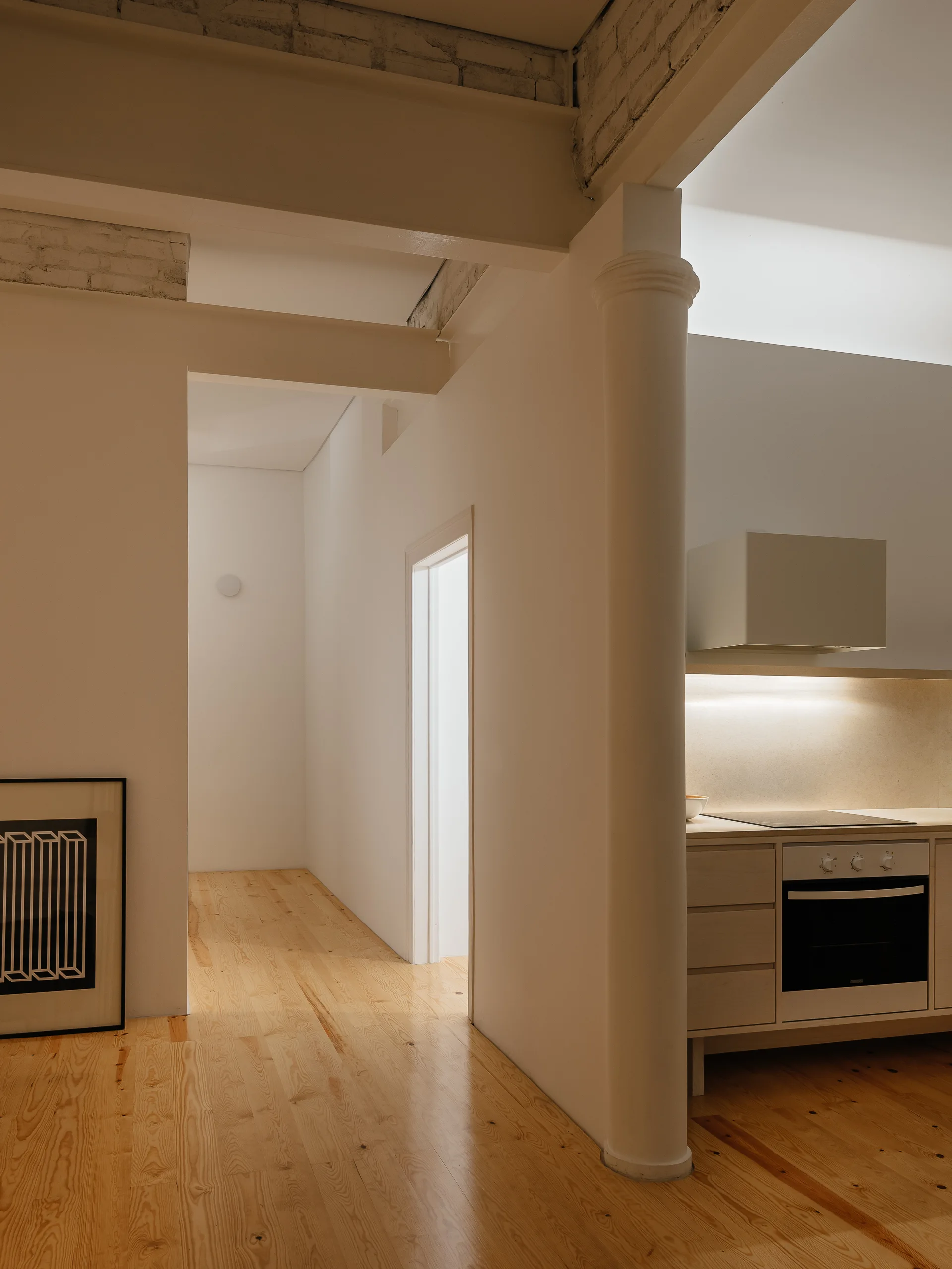 A minimalist hallway with light wood floors leading past a round white column to a modern kitchen with light wood cabinets and an oven.