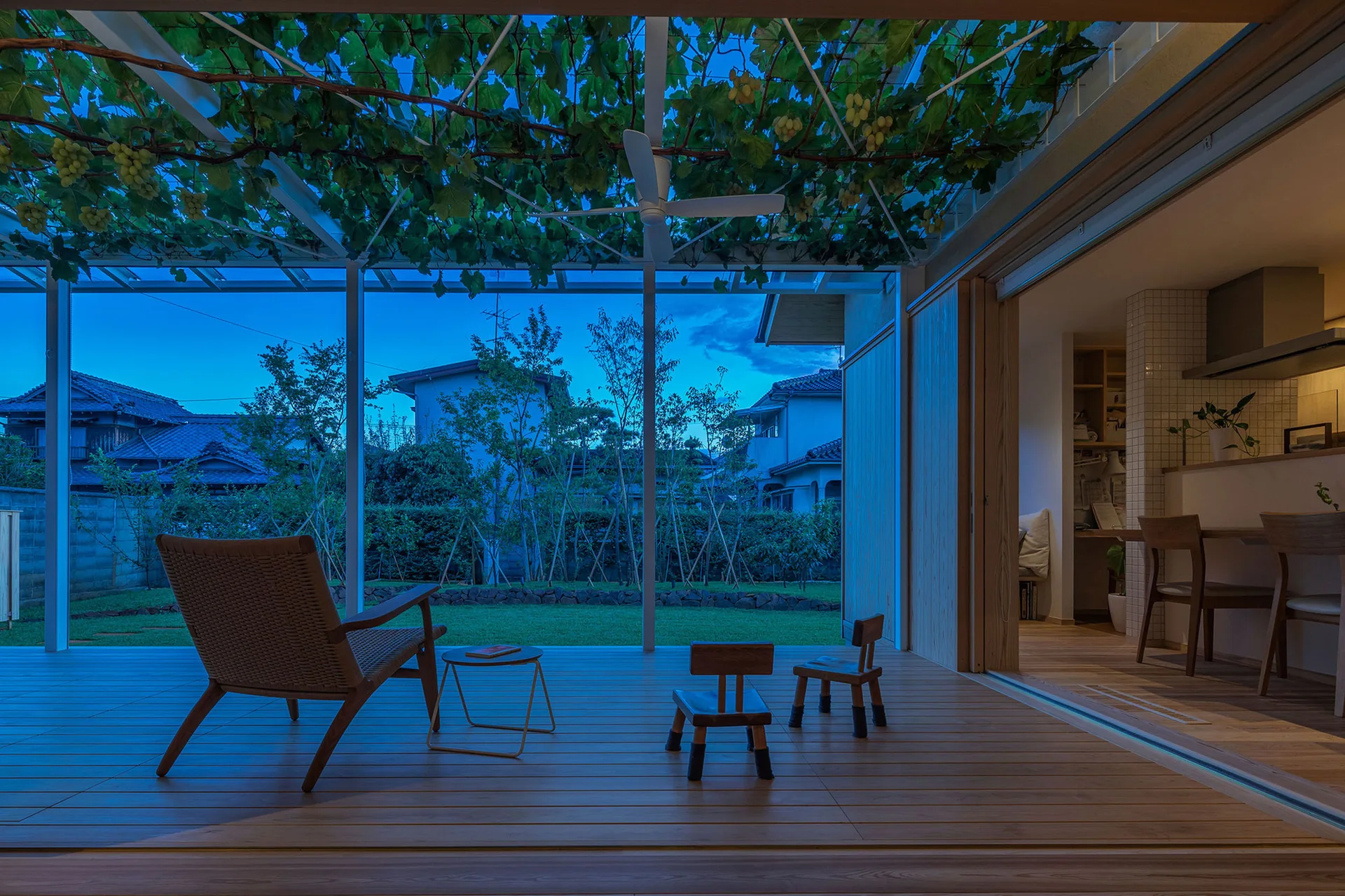A covered wooden deck at twilight features a woven lounge chair, small wooden stools, and an overhead pergola frame covered with leafy green grapevines.