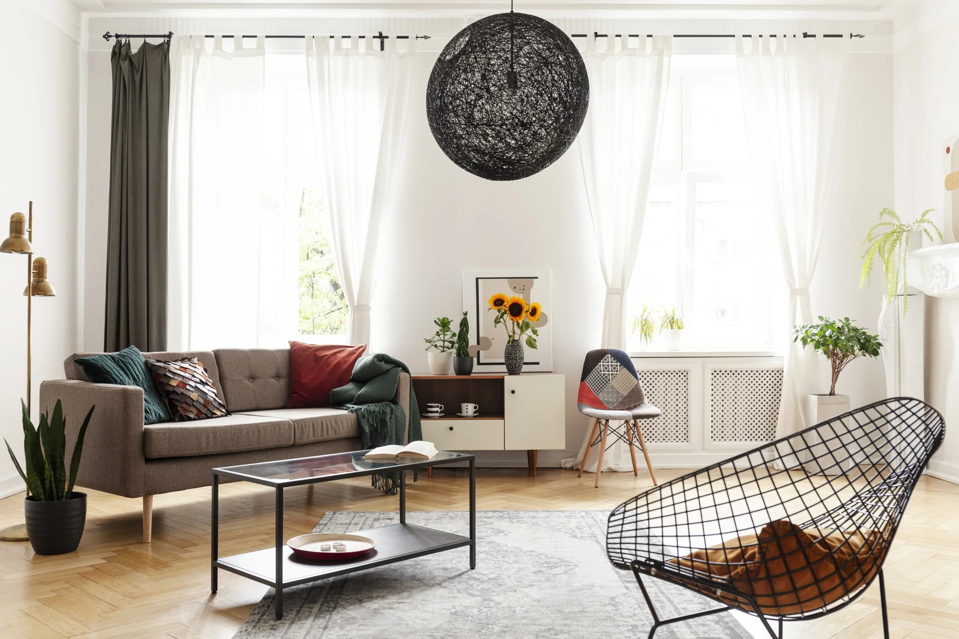 Bright living room with herringbone wood floors, a brown sofa, a glass coffee table, a black wire lounge chair, and a large black woven pendant light.