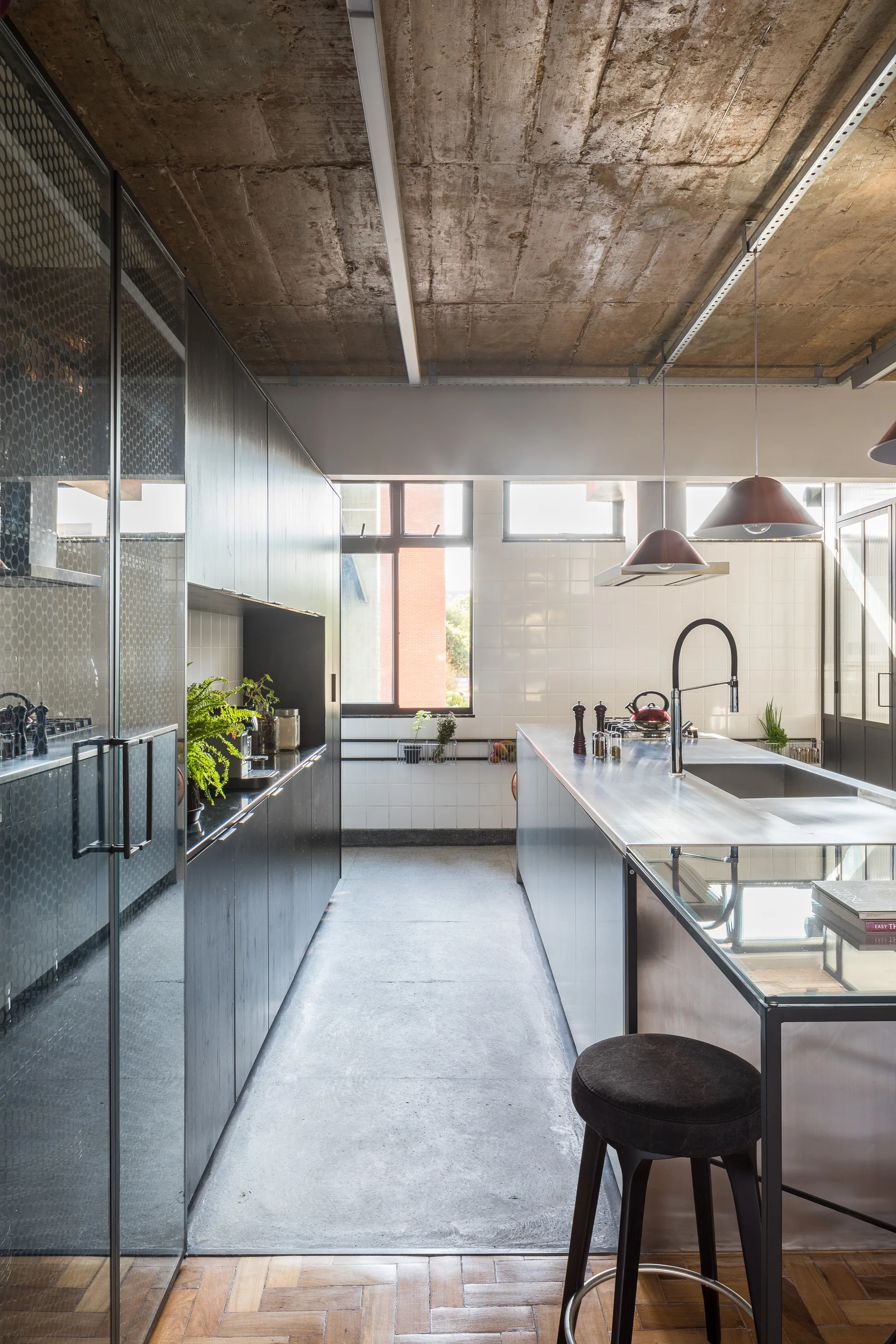 A modern kitchen with dark matte cabinetry, a stainless steel island, an exposed concrete ceiling, and a transition from parquet to concrete flooring.