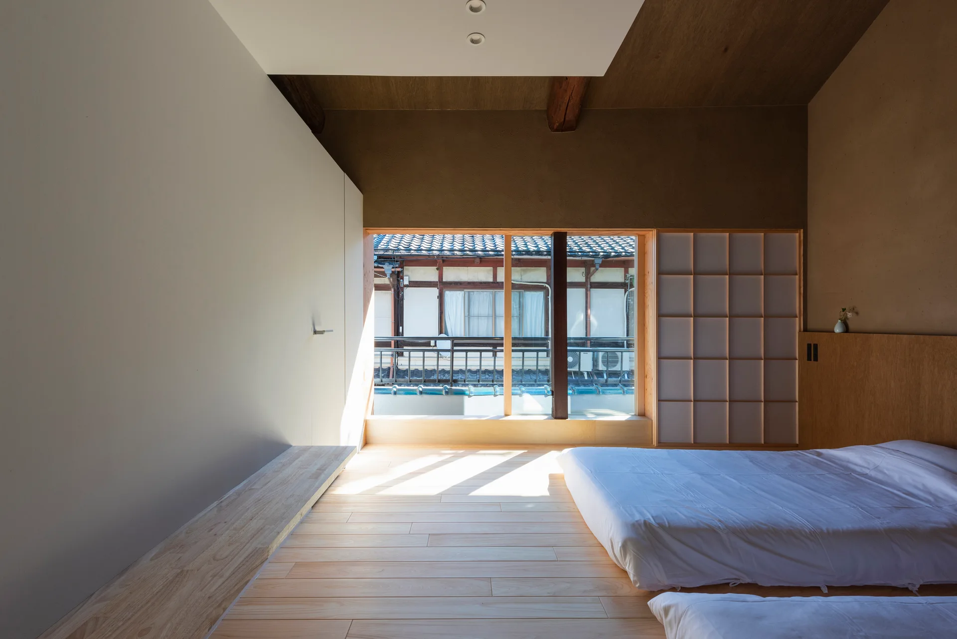 A minimalist bedroom features light wood plank flooring, a low-profile white bed, a grid-patterned shoji screen, and a large window.