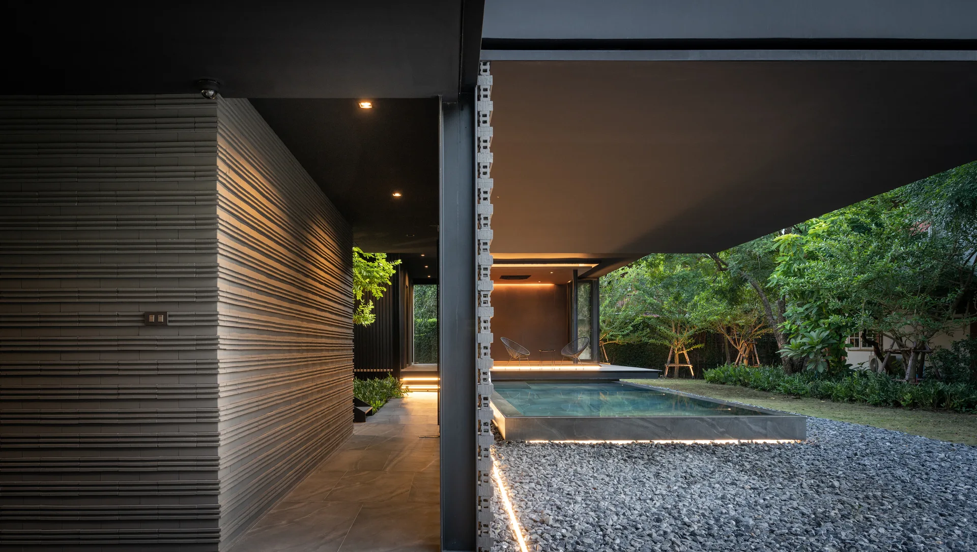 A modern dark grey patio features a rectangular swimming pool, gravel ground, a textured brick wall, and two wire chairs under a wide overhanging roof.