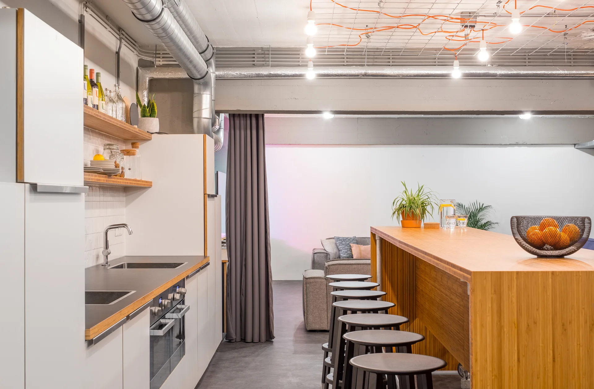 An industrial loft kitchen features a long wooden island with bar stools, white cabinetry, and exposed metal ductwork on a concrete ceiling.