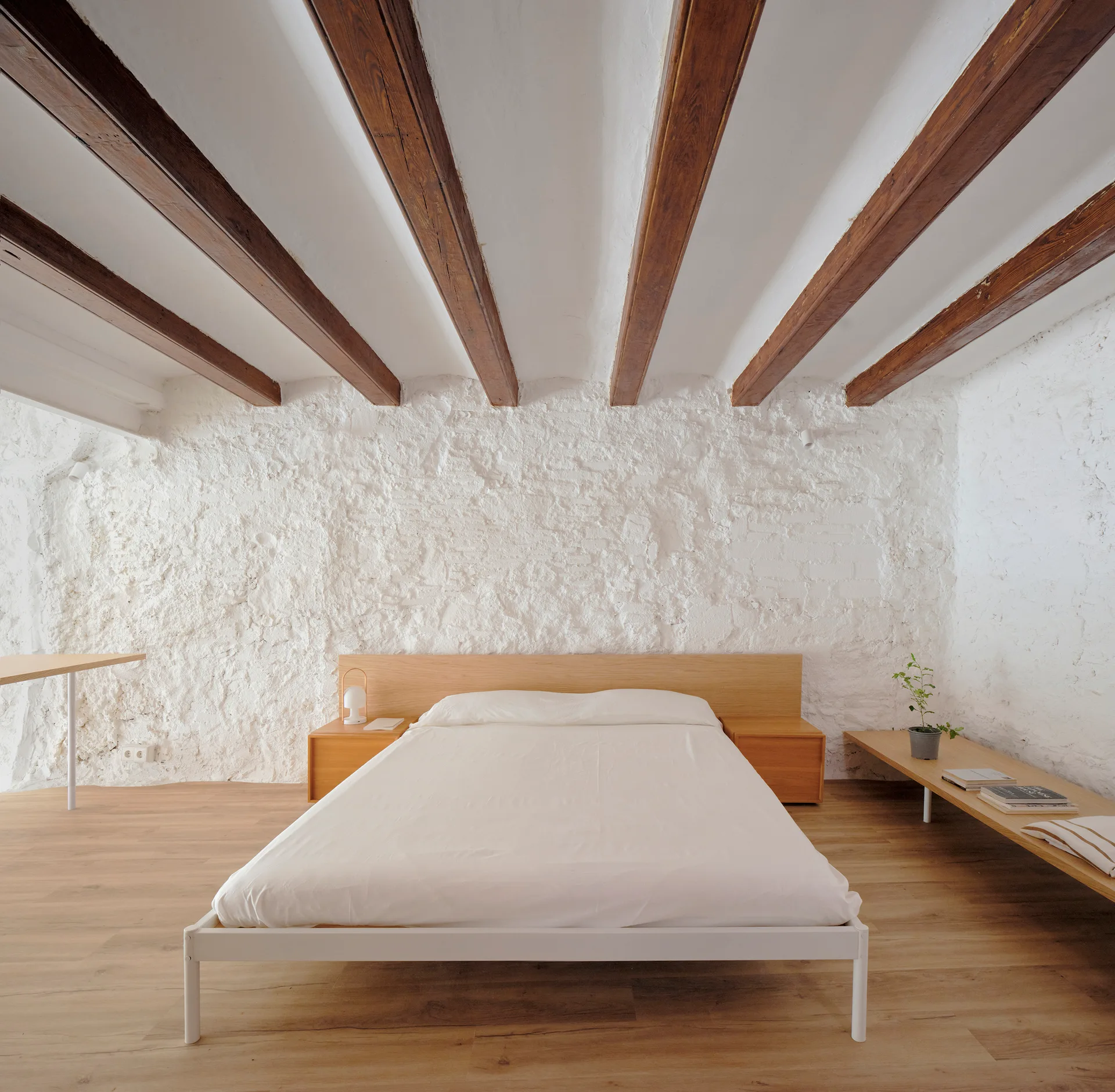 A minimalist bedroom features a white bed beneath exposed wood ceiling beams, backed by a highly textured white stone accent wall.
